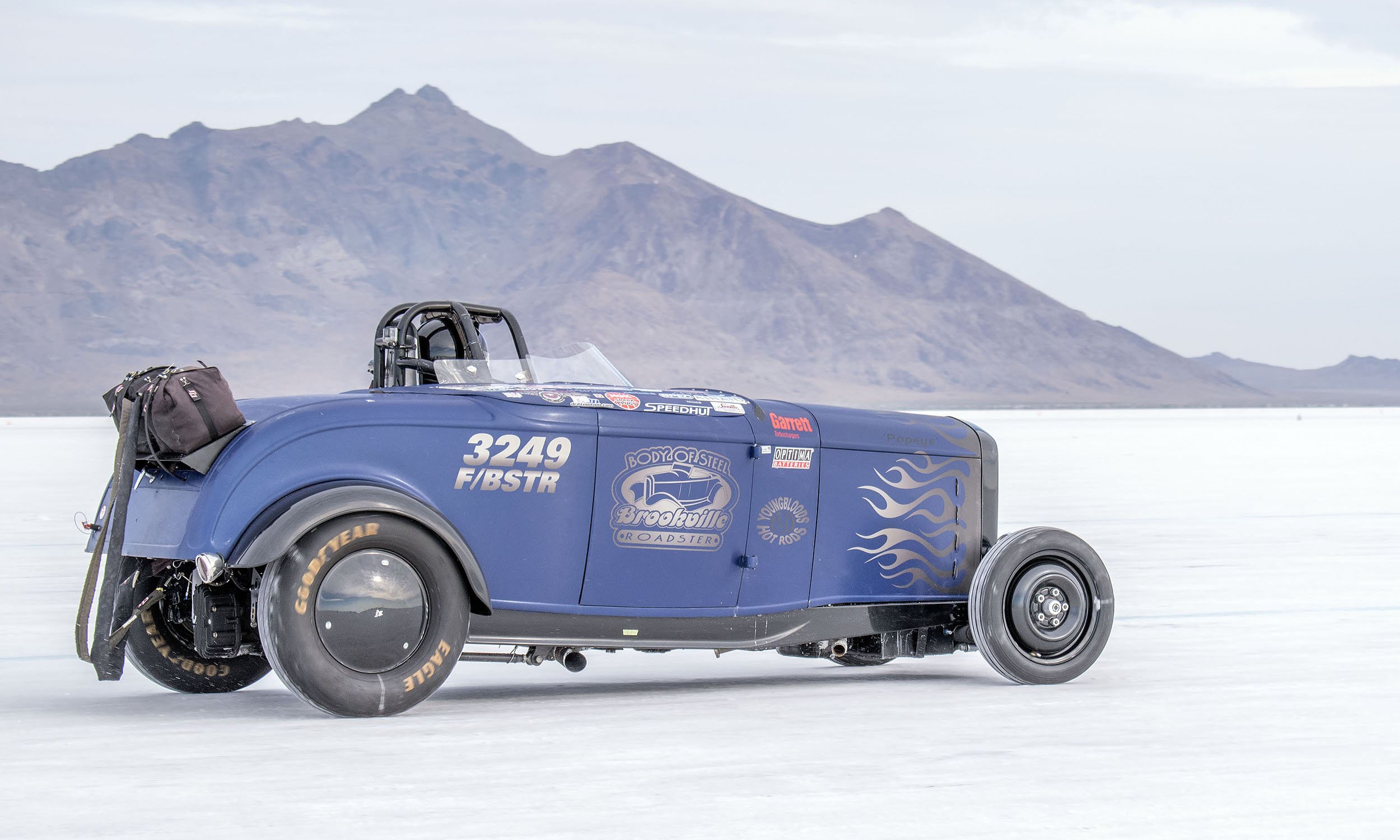 A blue land speed record car with racing decals, driving on a salt flat with mountains in the background.