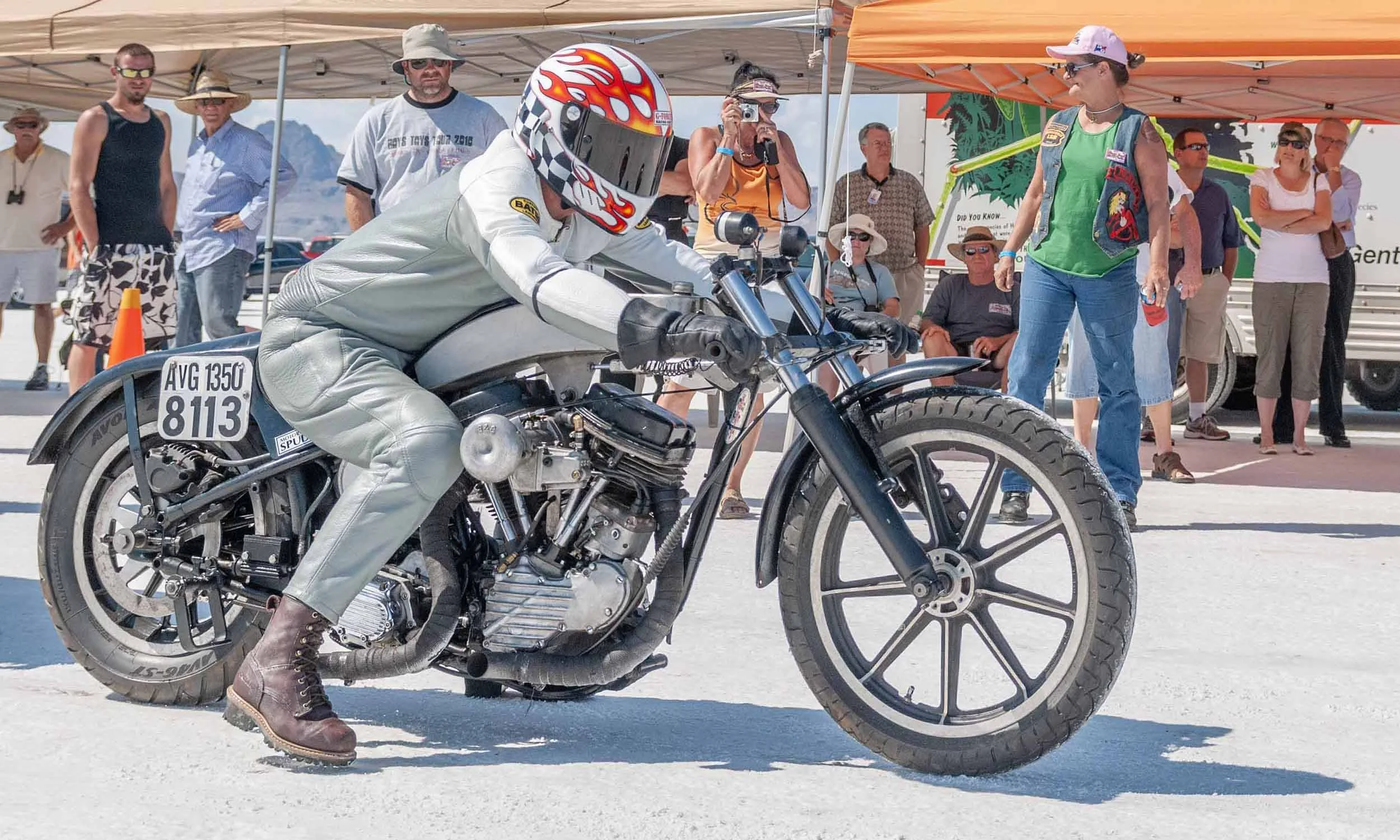 A person dressed in a silver racing suit and brown boots rides a vintage motorcycle at an outdoor event, surrounded by onlookers under tents.