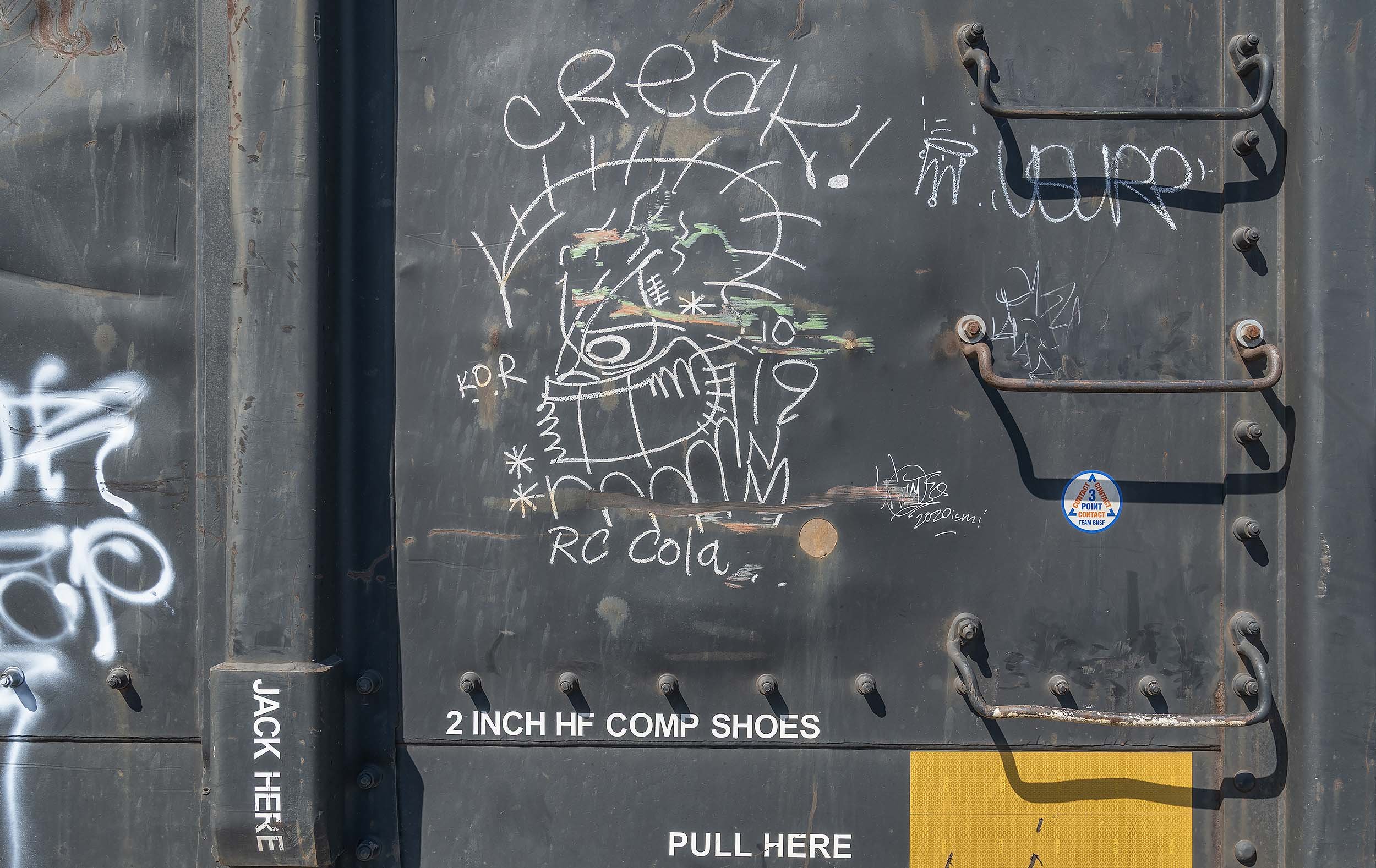 Graffiti and street art on a metal surface, including a colorful face or creature drawn with colored chalk, surrounded by various words and phrases like 'awesome' and 'RC cola', with a sticker and a handle for lifting or opening.