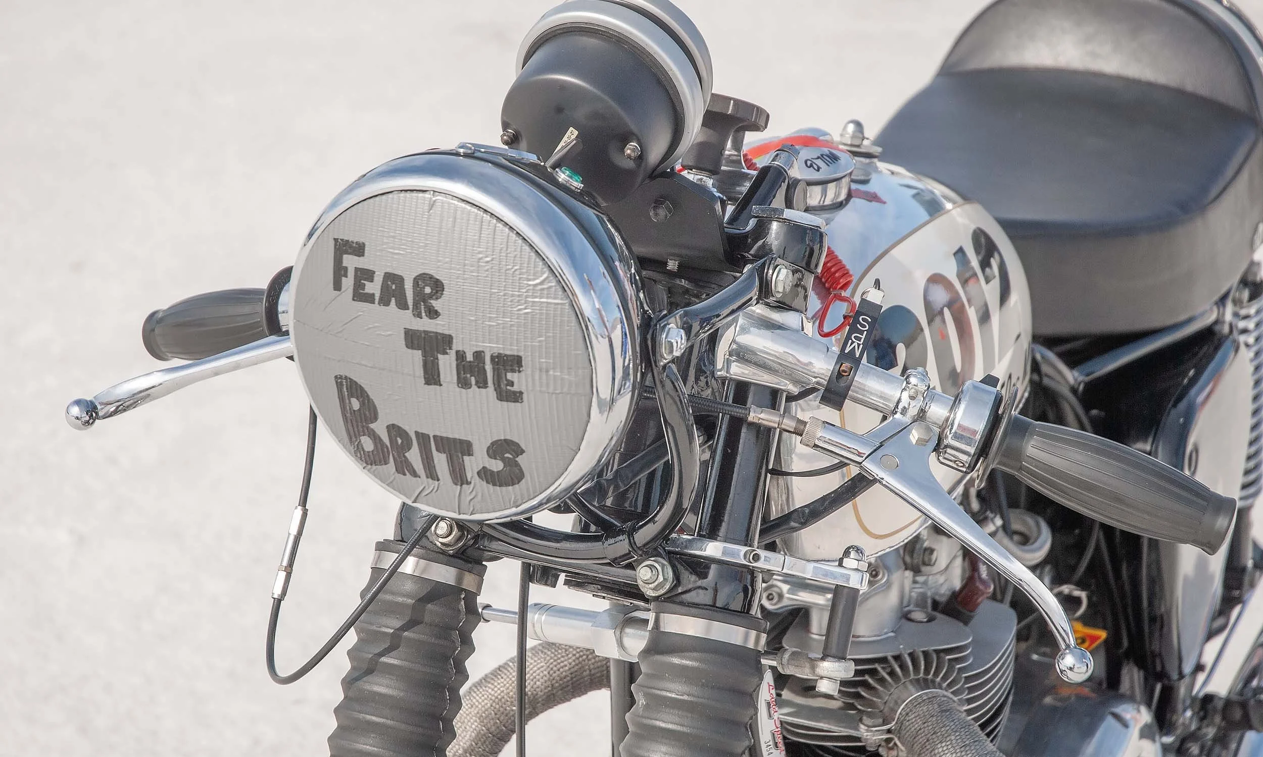 Close-up of a vintage  BSA motorcycle's front section, with a shiny metal headlight cover that reads 'FEAR THE BRITS,' Bonneville Salt Flat Racing