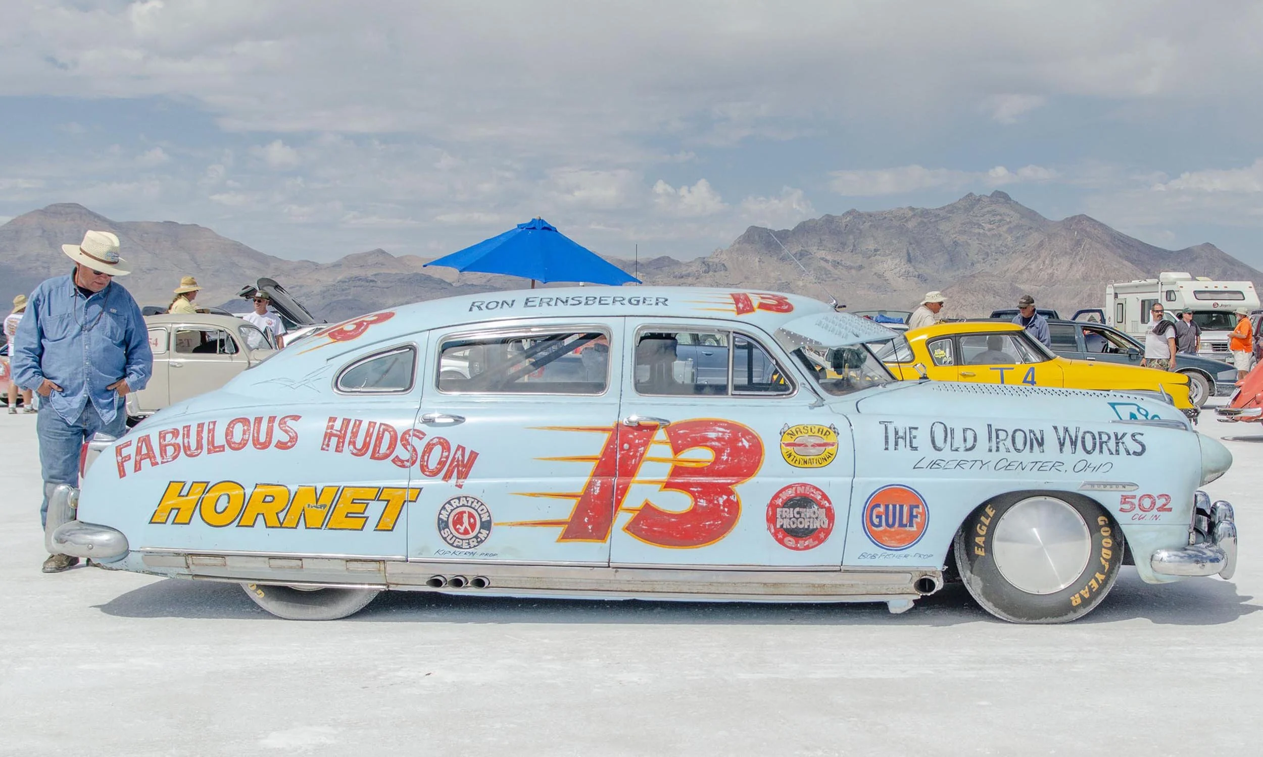 Vintage race car with various sponsor logos and the number 13, parked on a desert with mountains in the background, surrounded by people including a man in a blue shirt and hat.