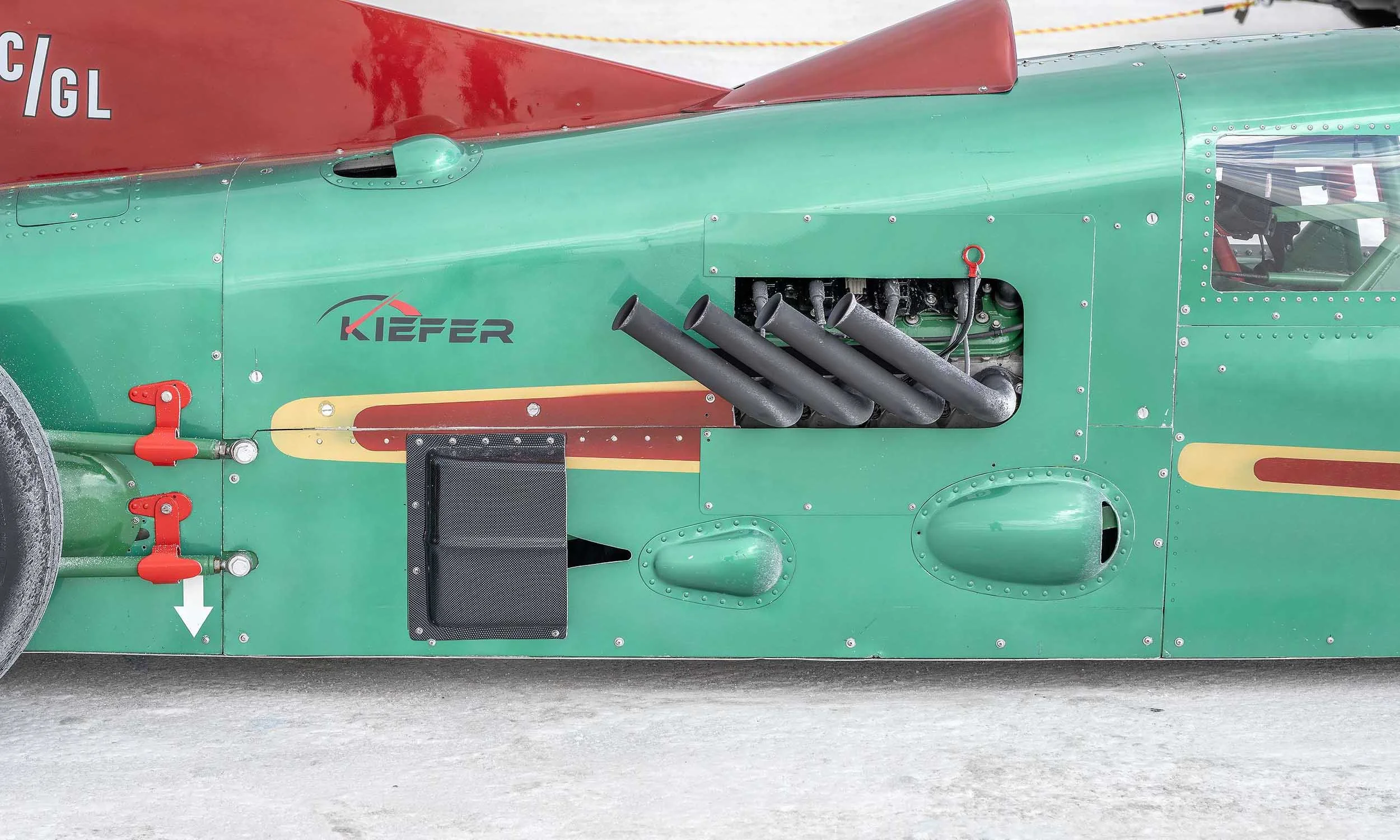 Close-up of a vintage race car with a green body, red accents, and the name 'Kiefer' painted on the side. The car features exhaust pipes and various mechanical components visible on the side.