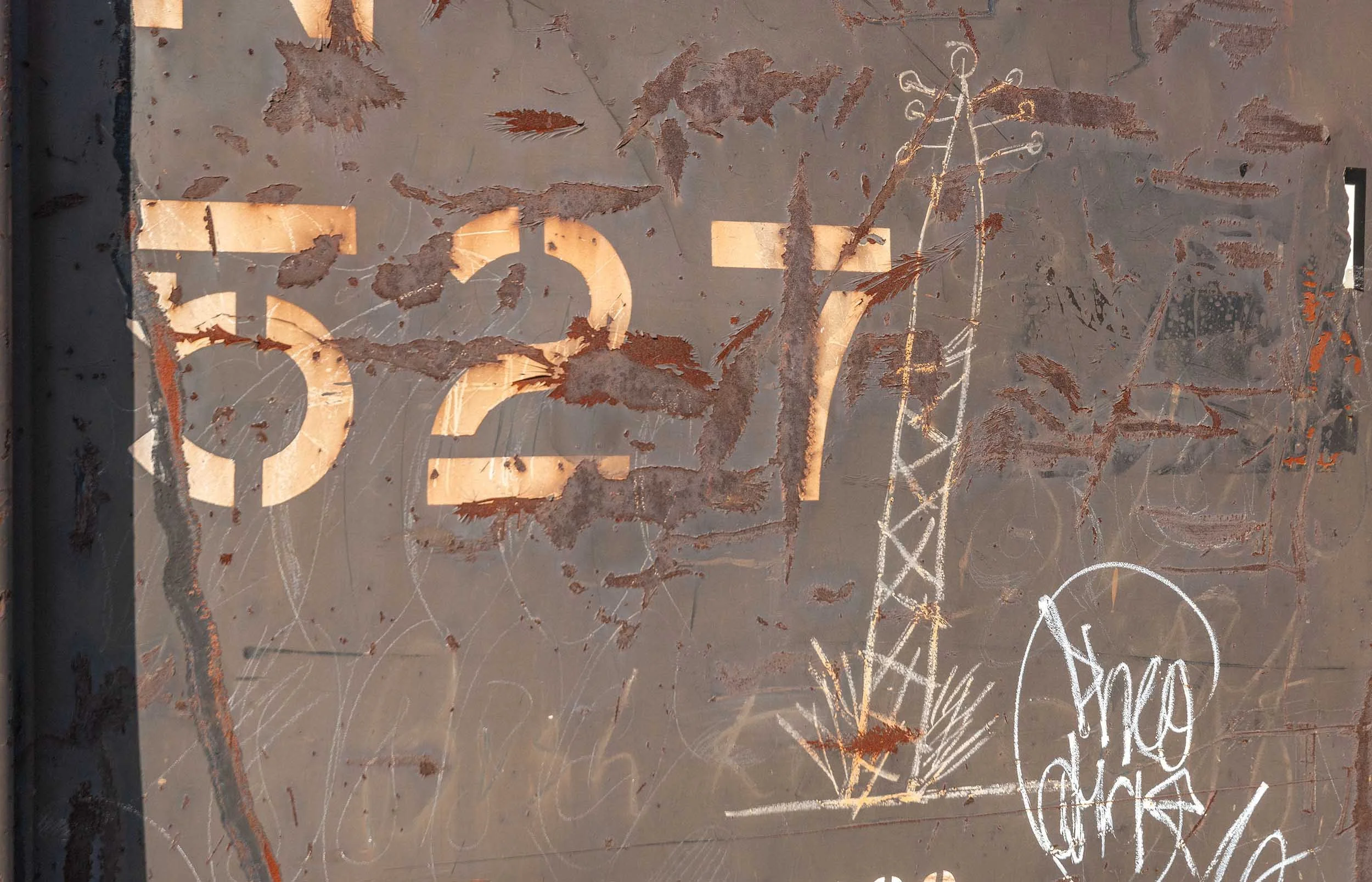 Rusty metal surface with faded white painted numbers and various scribbles, markings, and dried leaves scattered across it.