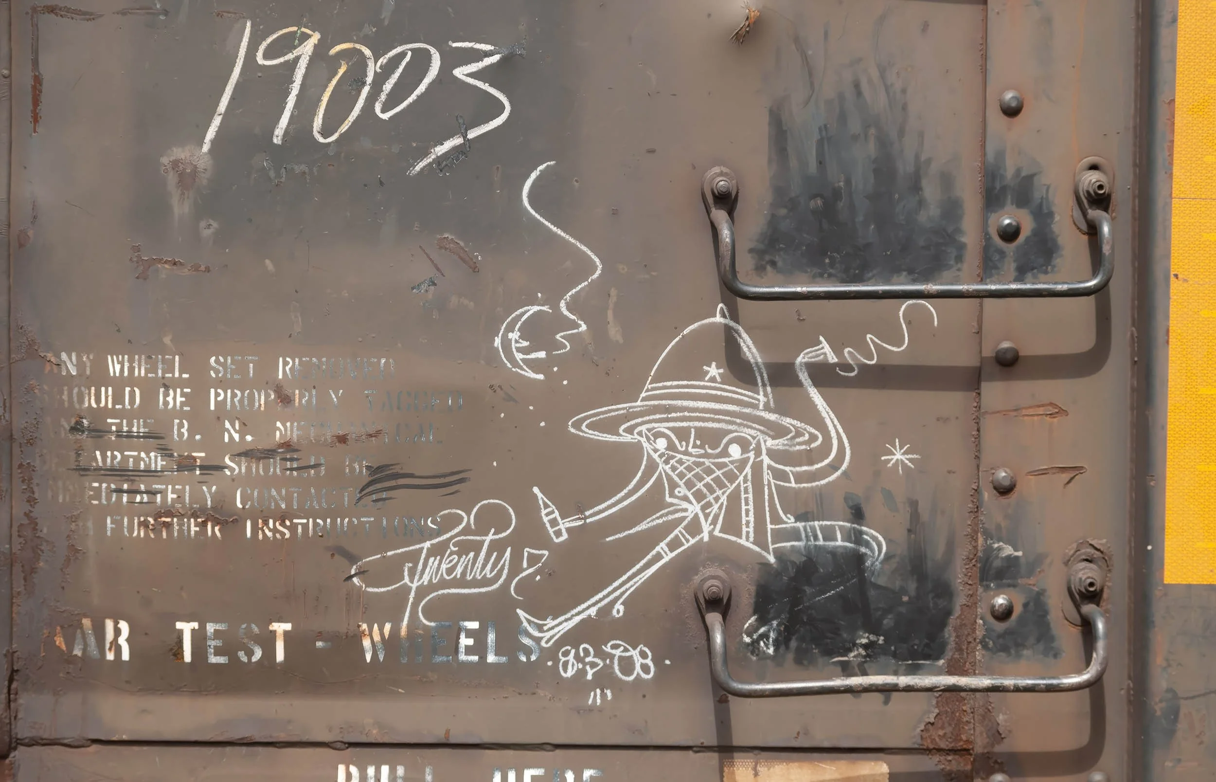 A rusty metal surface with various drawings and faded printed text. The drawings include a whimsical face with a hat and glasses, topped with a flying saucer, and stars. The text includes handwritten numbers '191005' and a phrase questioning if somet