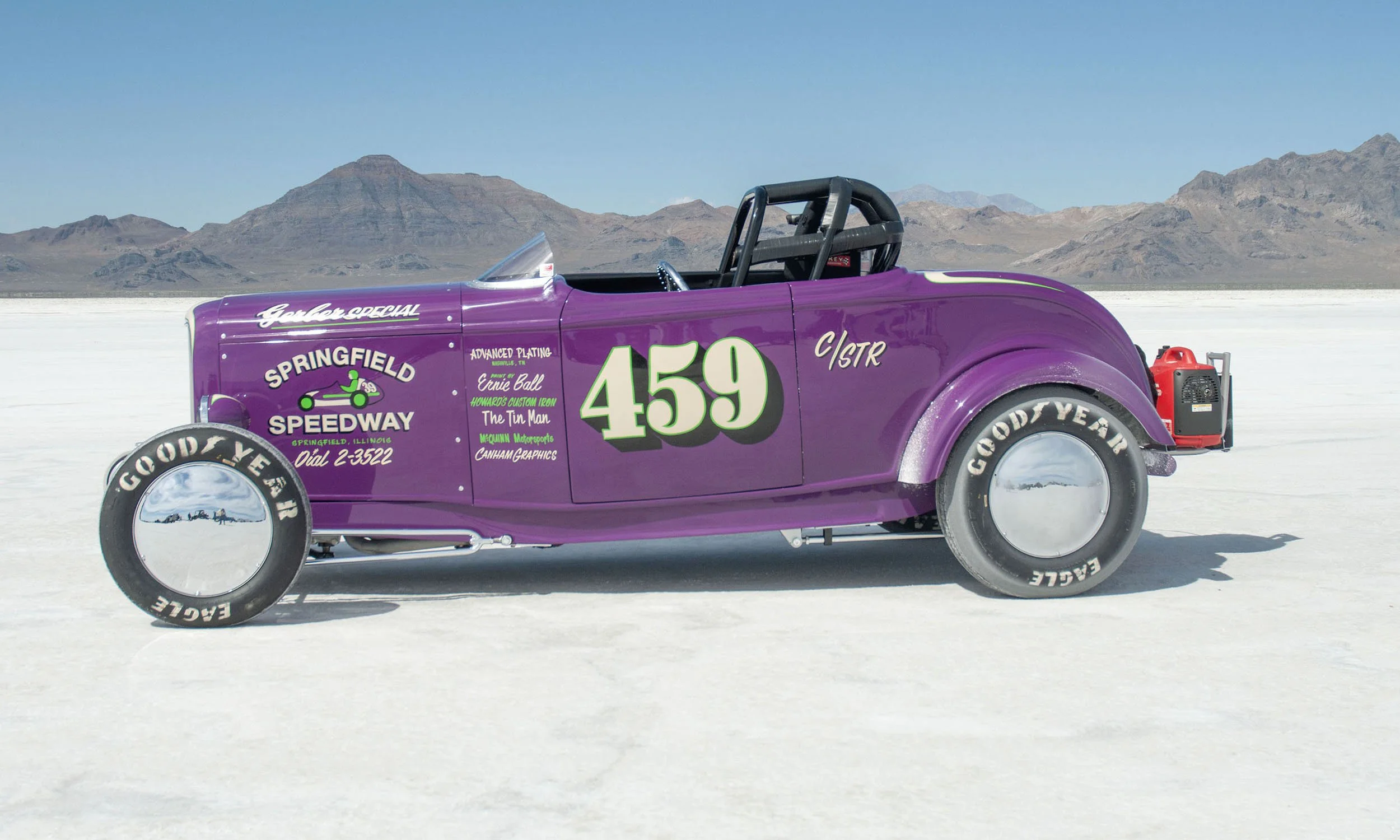 A vintage purple race car with the number 459 on the side, parked on a salt flat with mountains in the background.