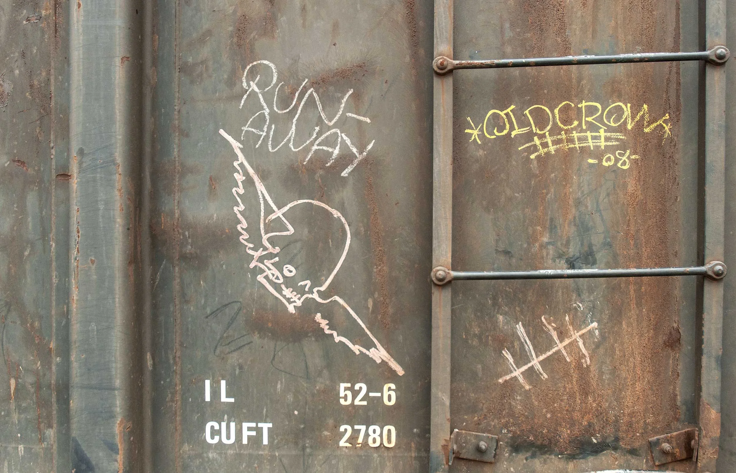 Rusty metal surface with chalk drawings of a hand holding a pencil, yellow graffiti reading 'OLD CROW' and some partially visible text, numbers, and symbols.