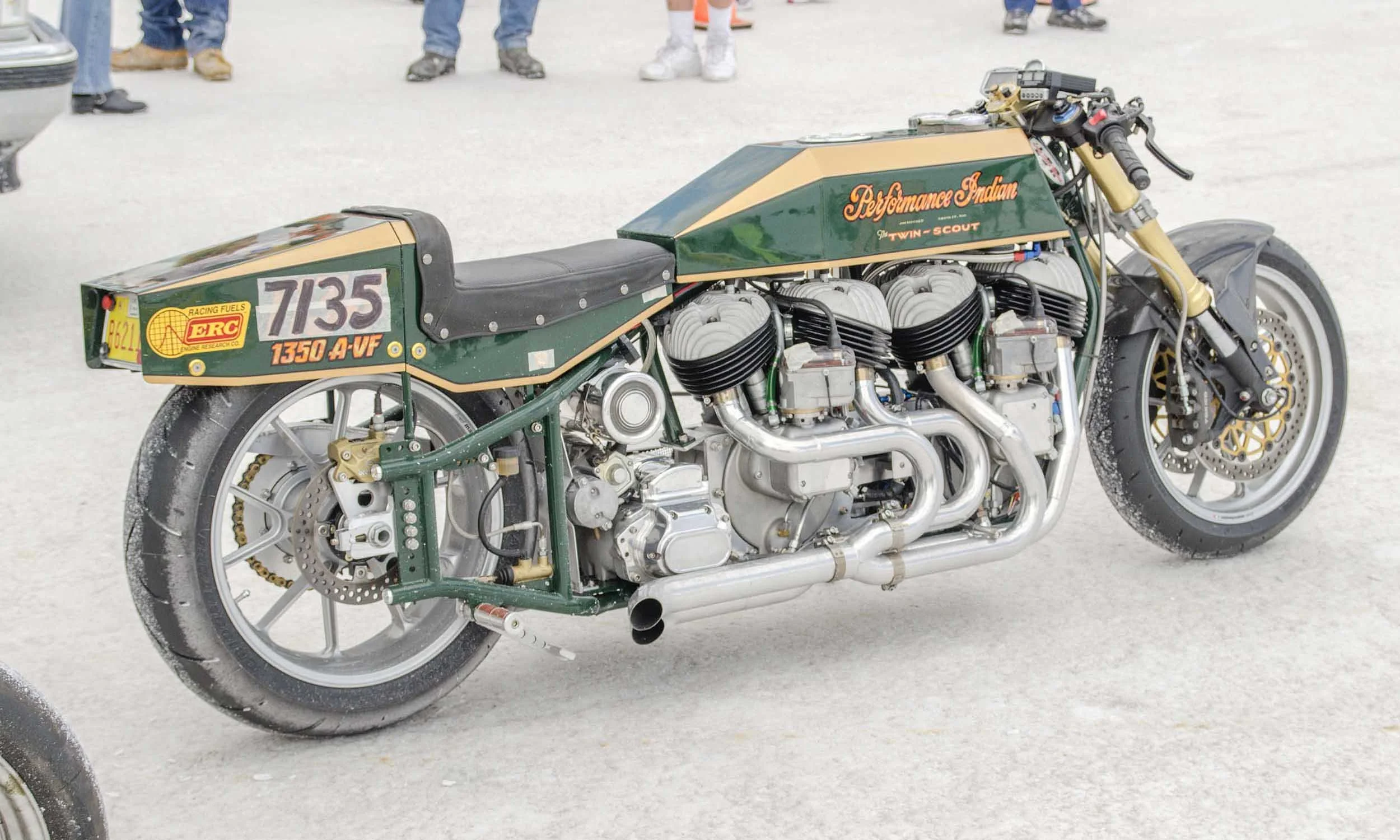 A vintage racing motorcycle with a green and gold color scheme, displaying the number 7135 and labeled as a Performance Indian Twin-Scout, parked on a salt flat with people in the background.