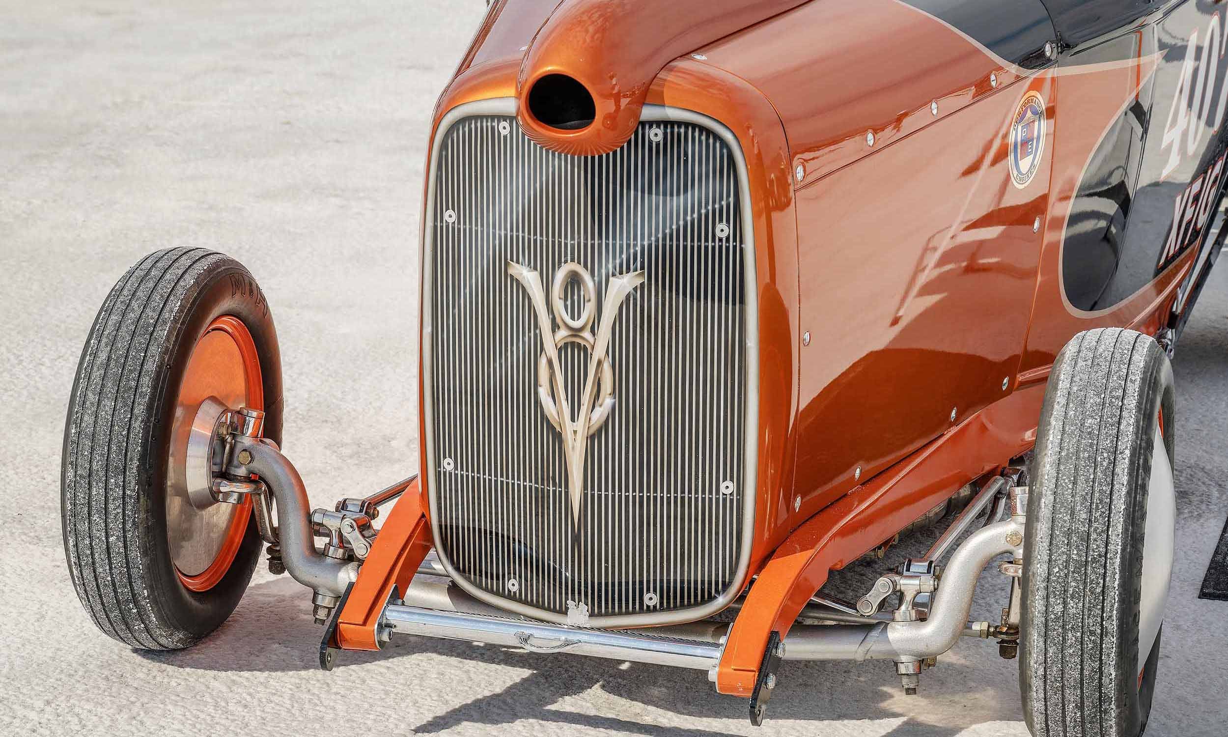 Close-up of a vintage race car with orange and black paint, silver grille, and prominent racing emblem.