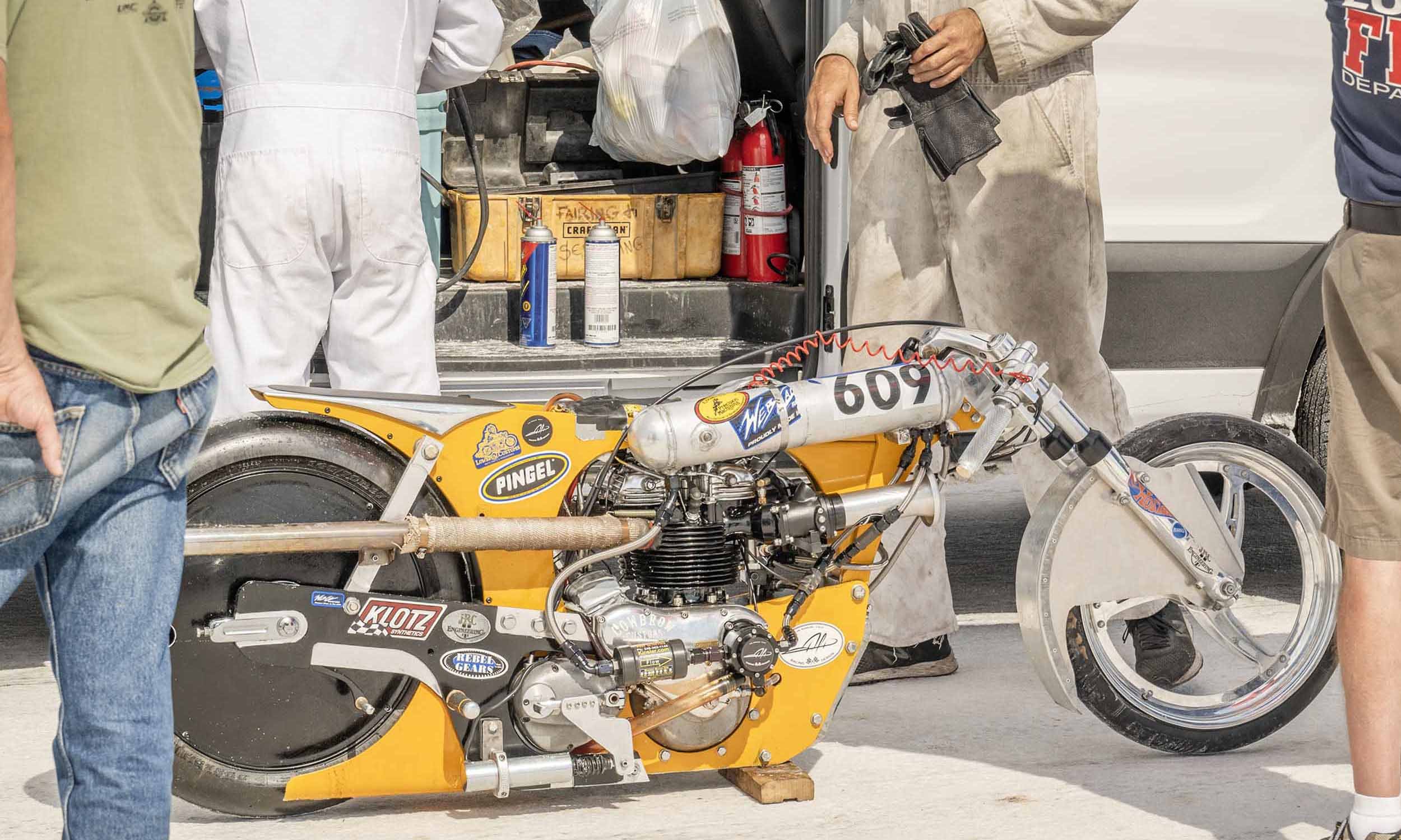 Yellow drag racing motorcycle without a rider, surrounded by people, in a pit area. The bike has various stickers and decals, and the engine is exposed.