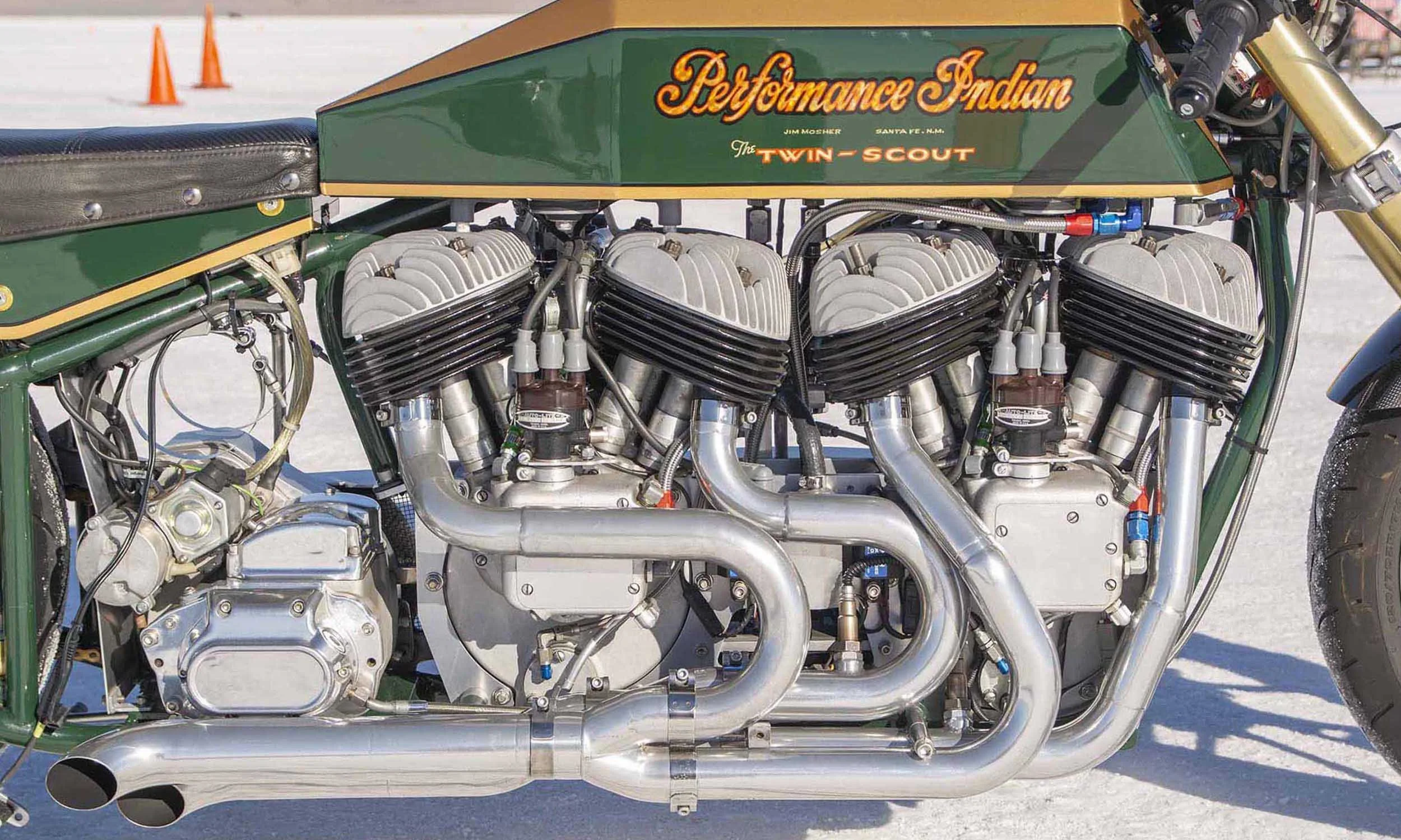 Close-up of a vintage race car engine with multiple cylinders, pipes, and mechanical components, with racing track and orange cones in the background.
