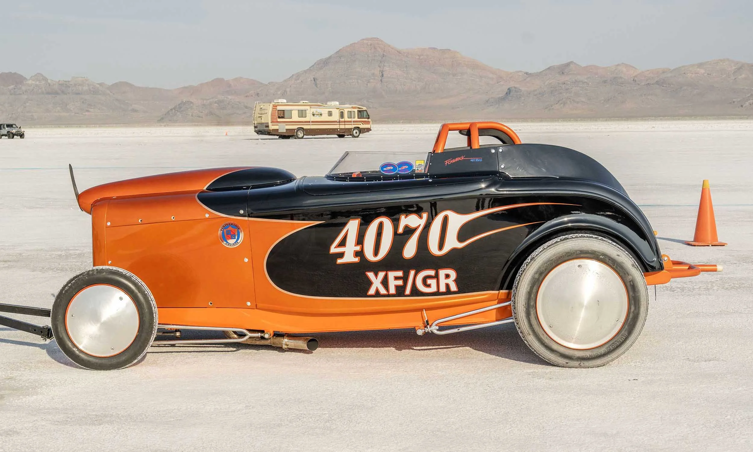 A vintage drag racing car with orange and black paint and the number 4070 on its side is on a salt flat track, with orange cones and distant mountains in the background.