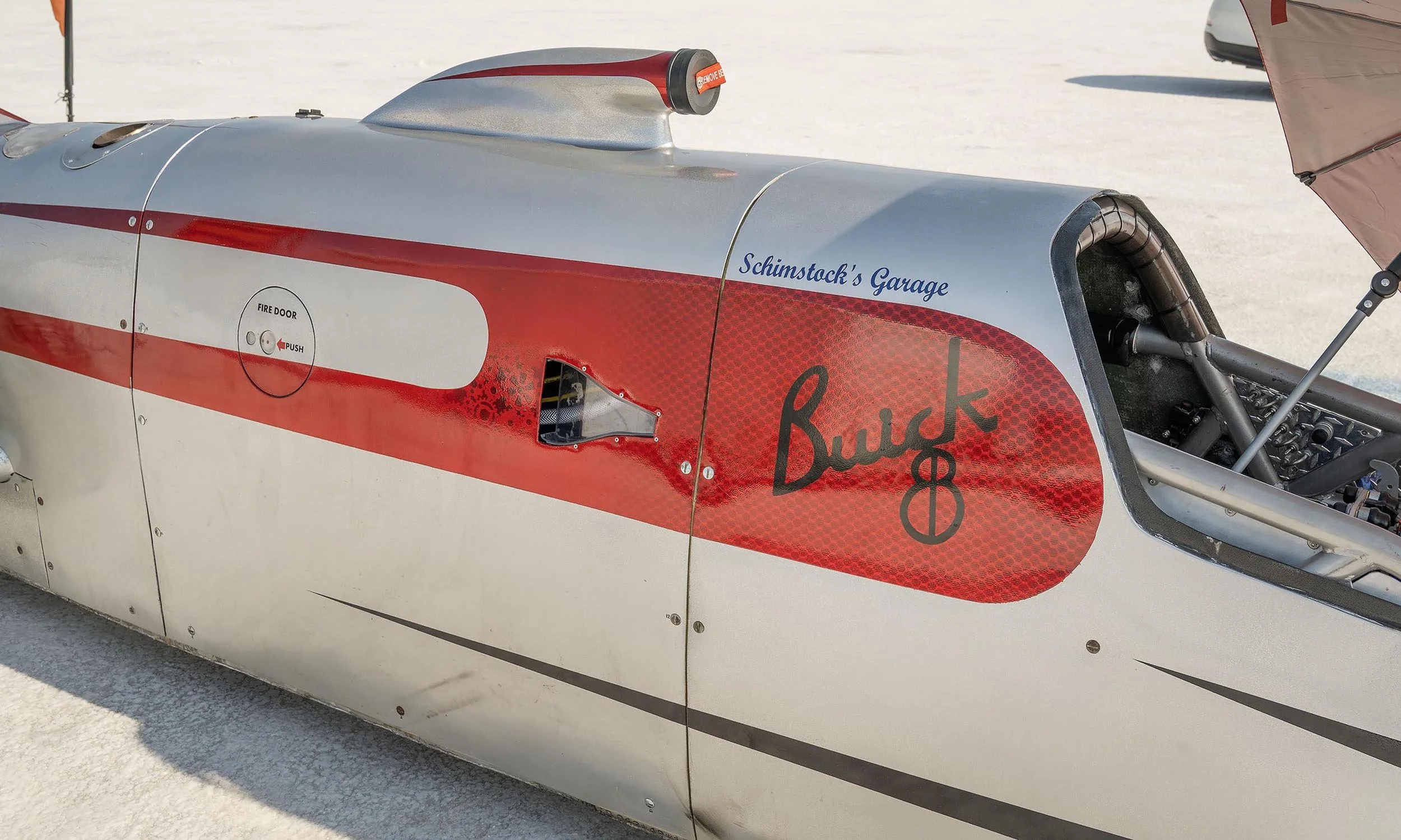 Close-up of a race car's side panel with a red and silver design, featuring signatures and sponsor logos, including one that reads 'Schimstock's Garage' and another that says 'Buck 8'.