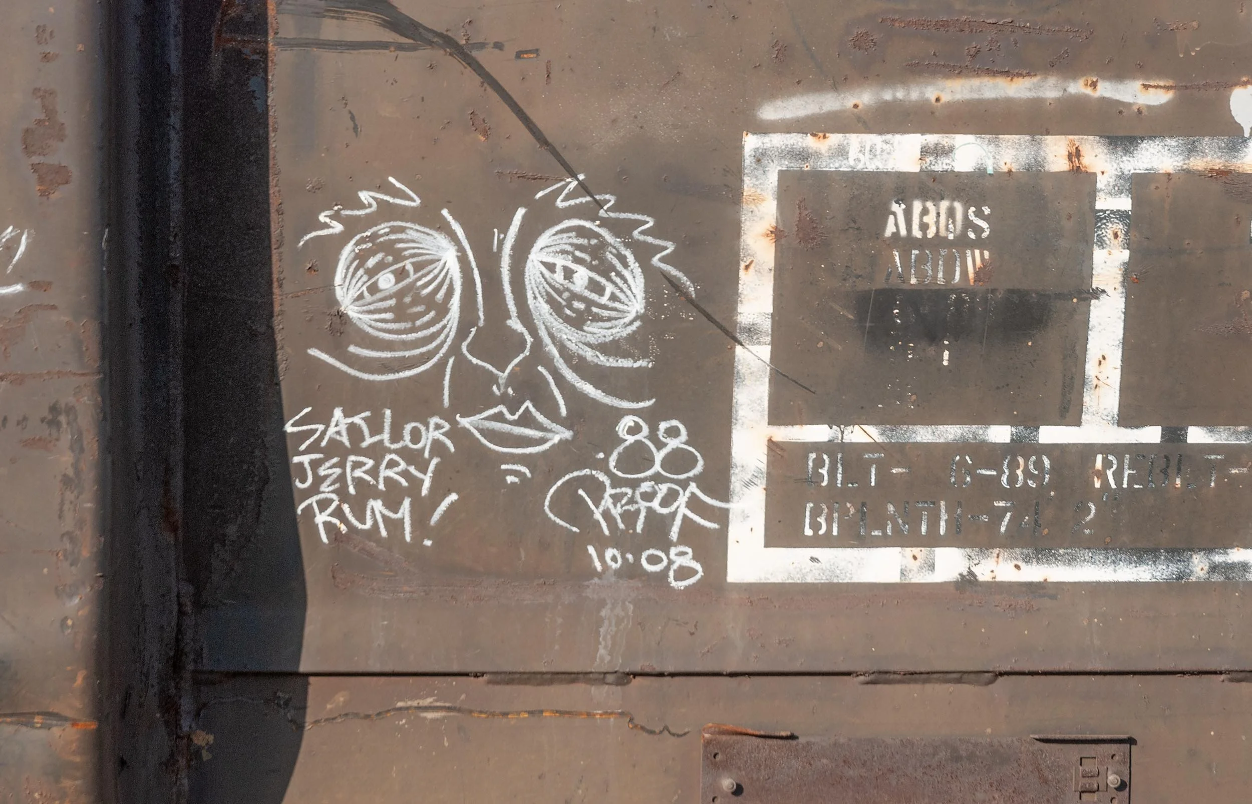 White chalk drawing of a face with sunglasses and hairstyle on a brown rusted metal surface, next to white stencil text and markings.