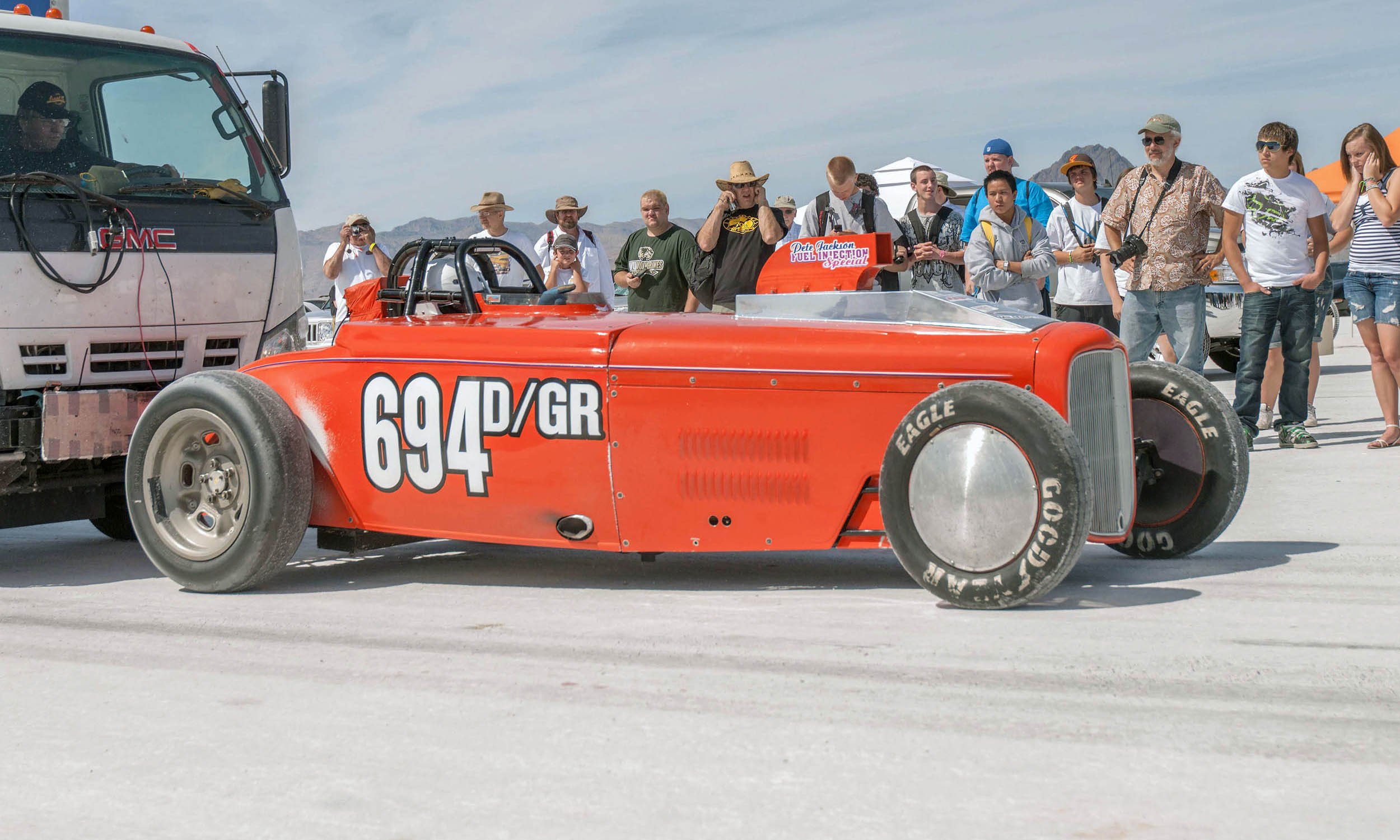 A vintage red race car with the number 694 and the words AD/GR on its side, parked on a dry salt flat. A group of people, some taking photos and others observing, stand behind the car. The background shows a mountain range and a clear blue sky.