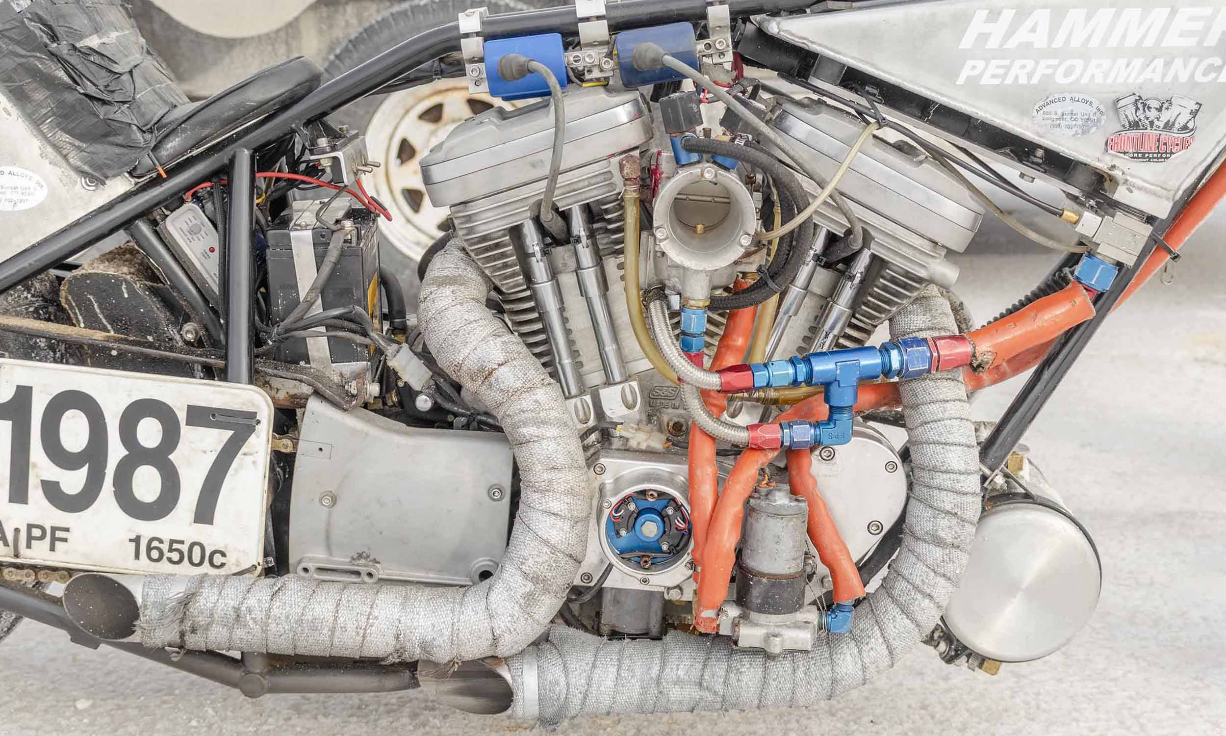 Close-up of a racing car engine with various pipes, wires, and components, including wrapped exhaust pipes and branding stickers.