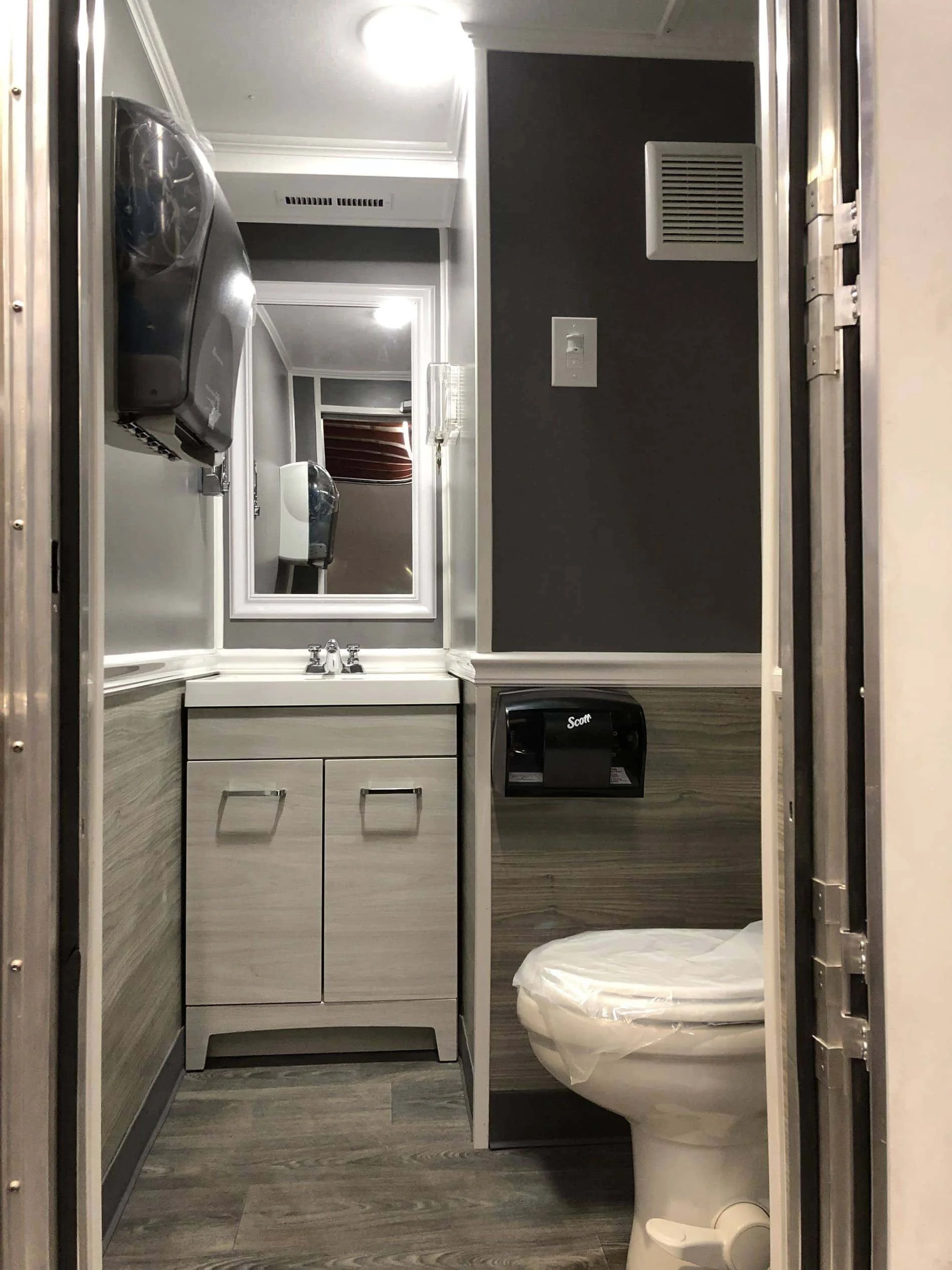 A small bathroom with a mirror, sink, cabinet, toilet, and wall vent. The toilet has a plastic cover, and there is a paper towel dispenser above the sink.