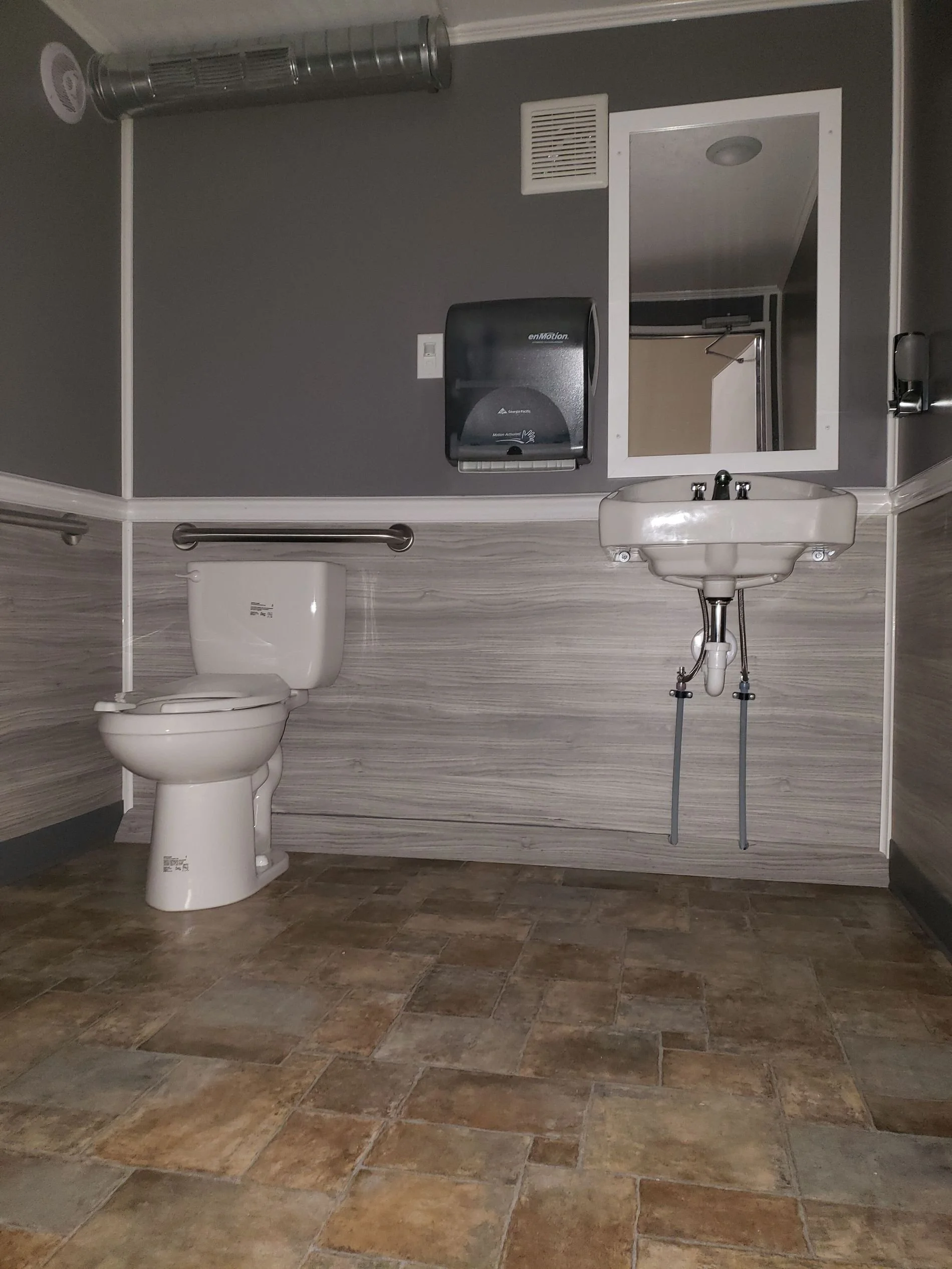 Bathroom with a toilet, a wall-mounted sink, a mirror, a hand sanitizer dispenser, and a hand dryer. The walls are gray and the floor has brown tiles.