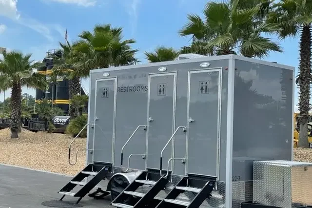 Portable restroom trailers with steps and handrails, set outdoors with palm trees in the background.