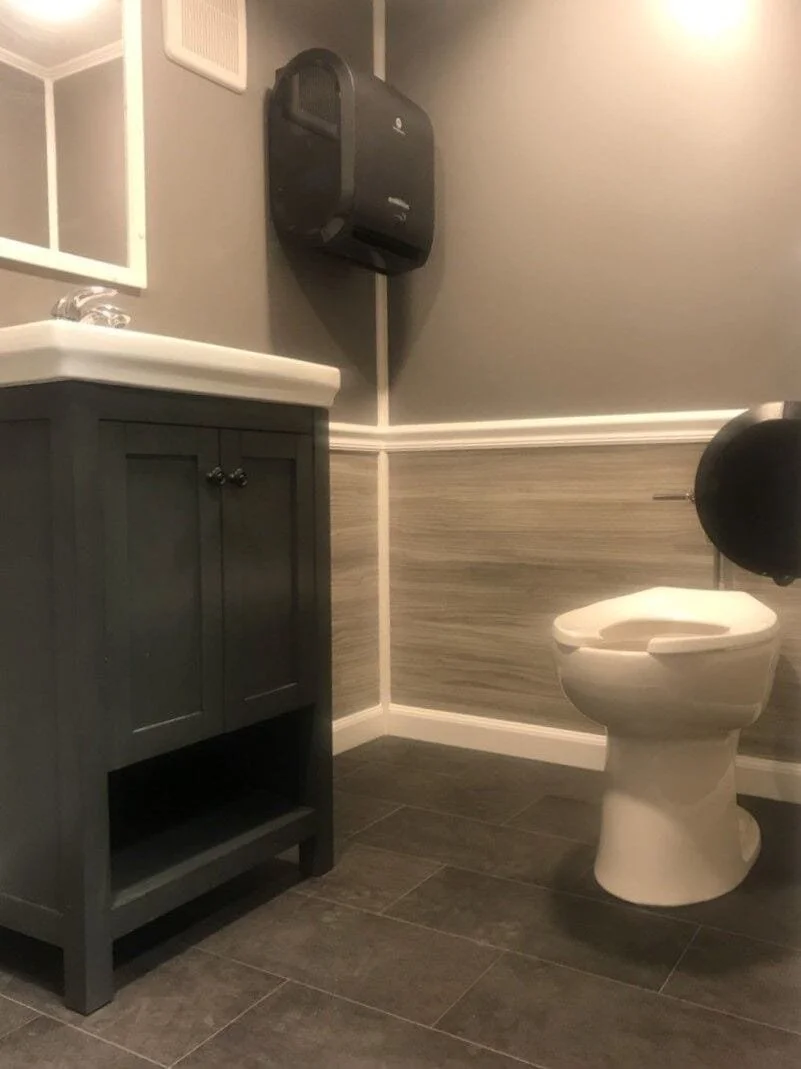 A bathroom with a gray vanity, a white sink, a toilet, a wall-mounted black paper towel dispenser, and gray and beige wainscoting on the walls.