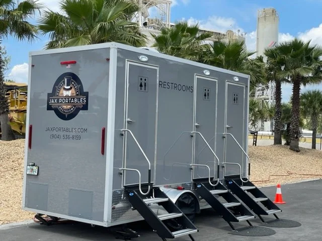 A portable restroom trailer with three separate stalls, labeled 'Restrooms,' positioned outdoors near palm trees and a parking area, with a logo for JAX Portables on the side.