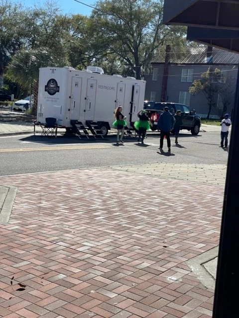 People standing outside a portable restroom trailer on a street, some dressed in green tutus. A building and parked cars are visible in the background.