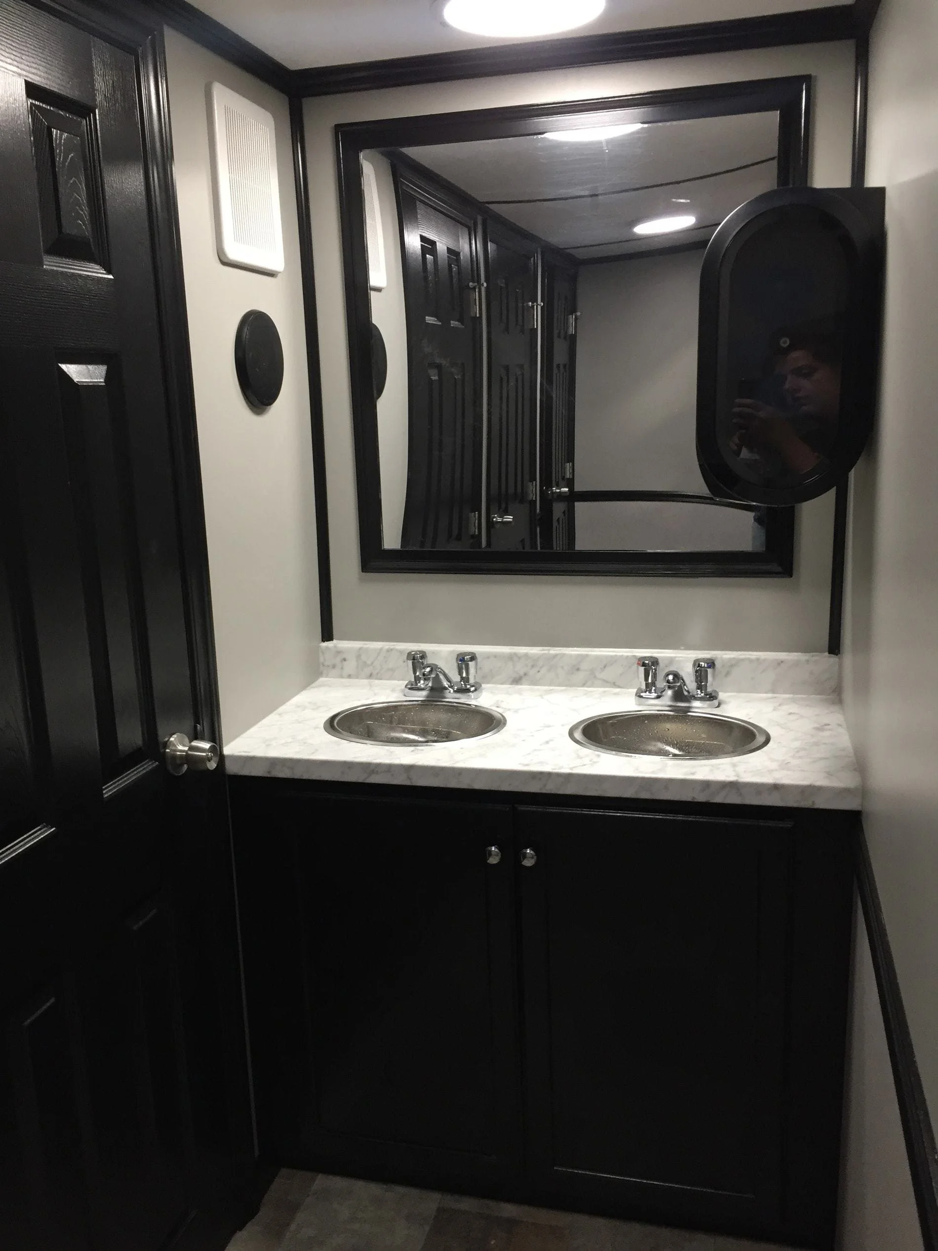 A bathroom vanity with two sinks, a marble countertop, a large mirror, and black cabinetry, with a woman taking a selfie visible in the mirror.