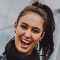 A young woman with dark hair pulled back in a high ponytail, smiling and showing her teeth, wearing a dark jacket.