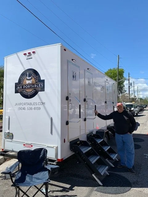 A man standing outside a large white portable restroom trailer with steps, logo, and website for Jax Portables, parked on a street with utility poles and cars in the background.