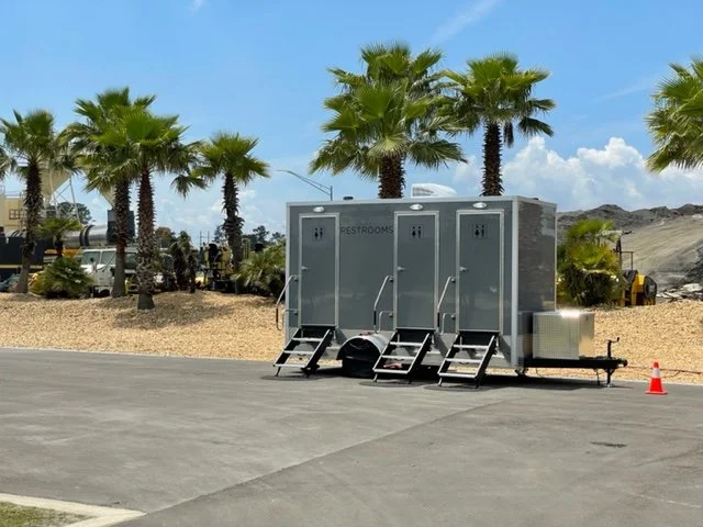 Portable restroom units on a paved surface with palm trees and construction equipment in the background.