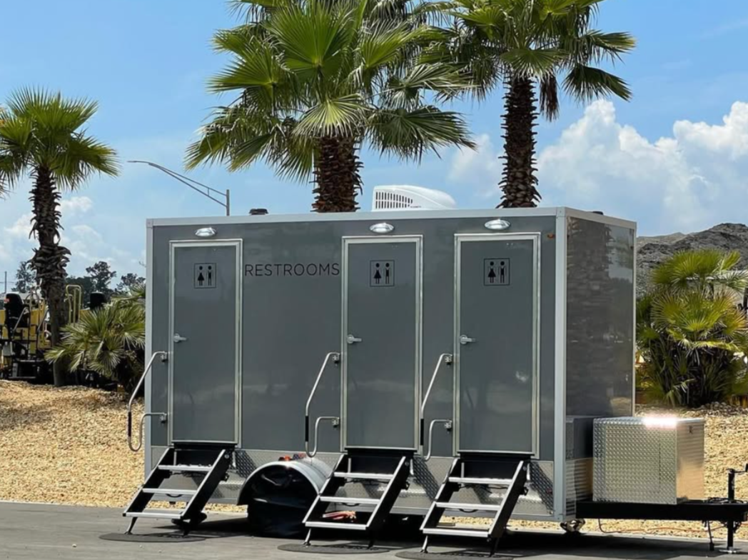 Portable restroom trailers with three doors and stairs, set outdoors with palm trees and a cloudy sky in the background.