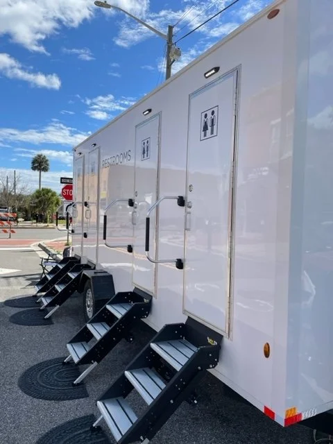 Portable restroom trailer with stairs and two doors, one marked for men and women, on paved outdoor area under a partly cloudy blue sky.
