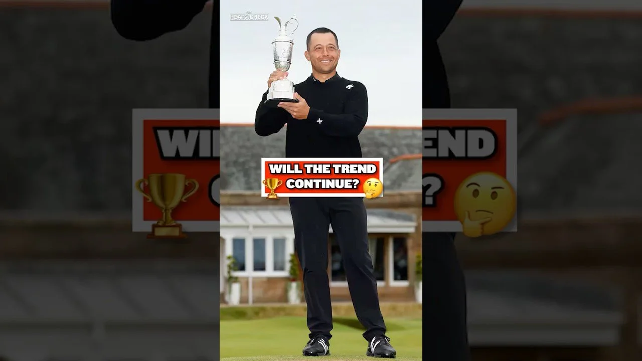 A man standing outdoors on a golf course, holding a trophy and smiling. He is dressed in a black jacket and pants. There are buildings in the background.