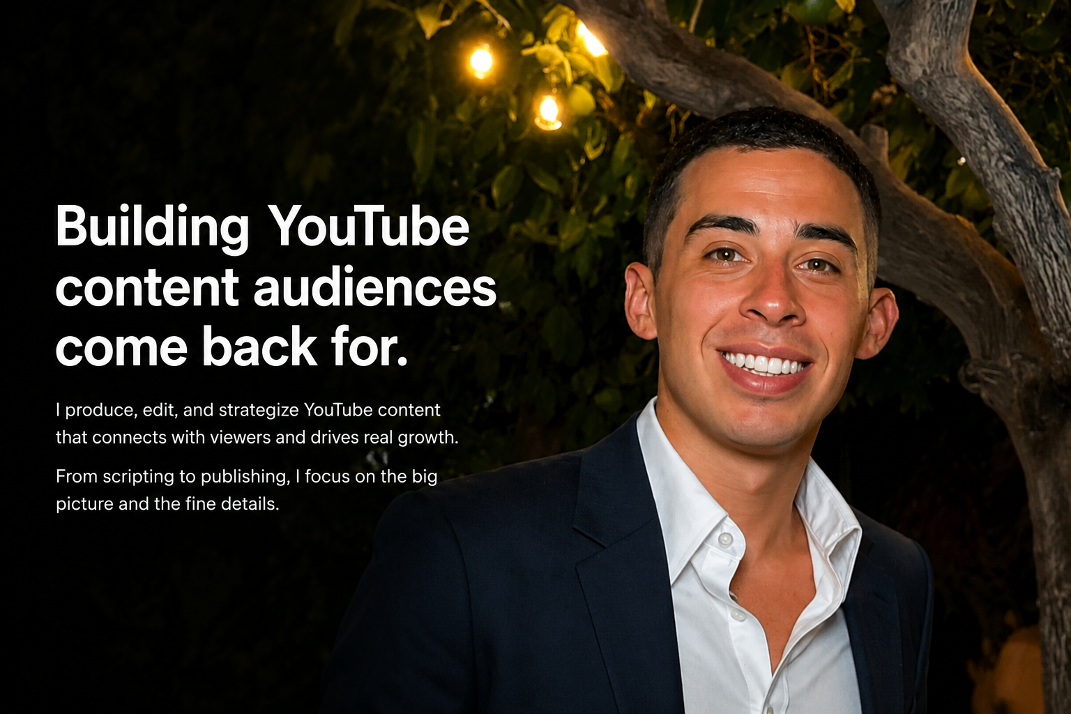 A smiling man in a suit with a white shirt under a dark blazer, standing outdoors at night with string lights and a tree in the background.