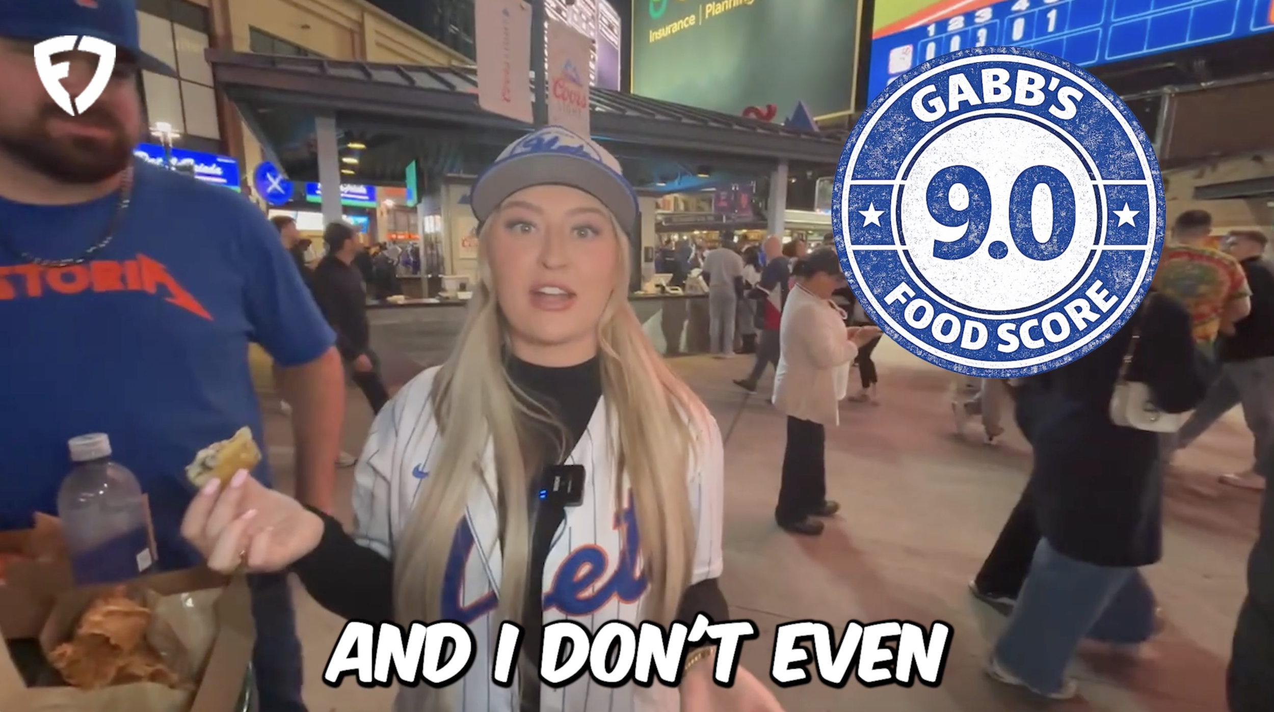 A young woman wearing a New York Mets jacket and standing in a busy outdoor area, possibly at night, with many people walking around and large digital screens in the background. There is a blue and white Gabb's Food Score badge on the right side of the image. The woman appears to be talking, and there are text overlays on the image that say 'AND I DON'T EVEN'.