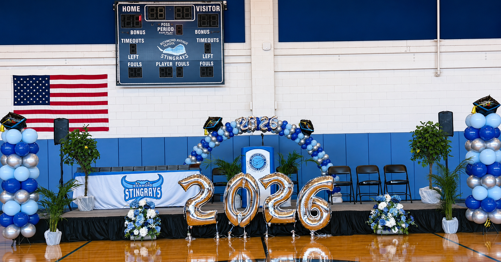 Indoor gymnasium decorated for a graduation or celebration with large balloons forming the year 2026, blue and silver balloons, a stage with a Richmond Stingrays logo, potted plants, chairs, and an American flag, with balloon bouquets and graduation caps.