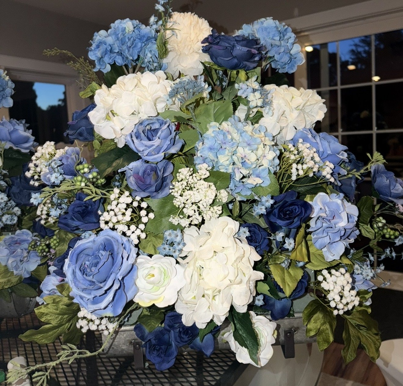 A large floral arrangement featuring white, light blue, and dark blue hydrangeas, roses, and other small white flowers with green leaves, placed indoors near a window.