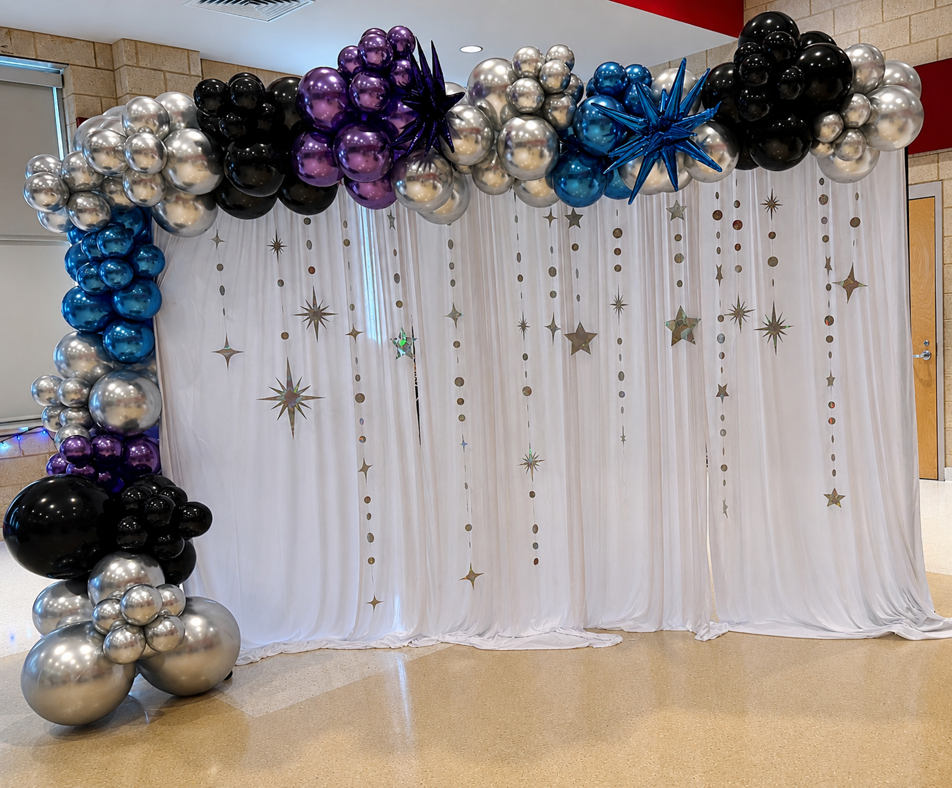 Decorative balloon arch with black, silver, purple, and blue balloons over white curtain with star and circle cutouts.