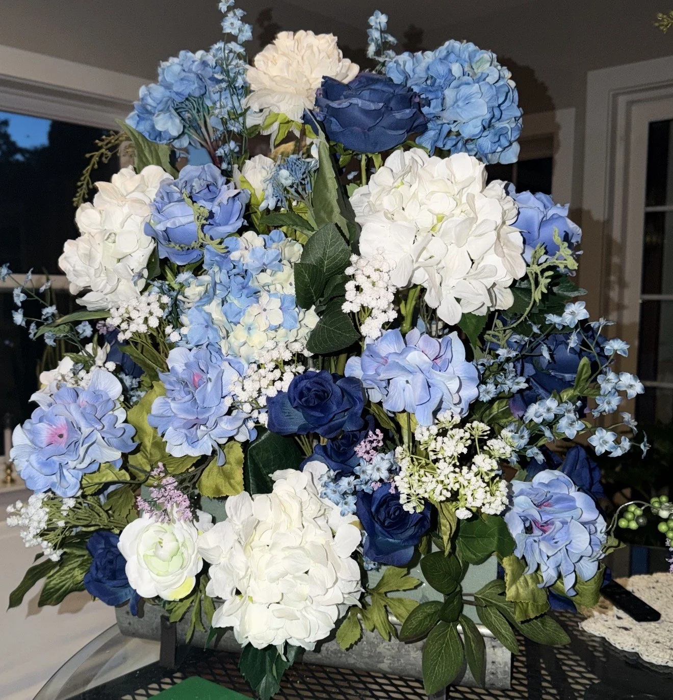 A bouquet of artificial blue and white hydrangeas, roses, and greenery in a container on a table near a window.