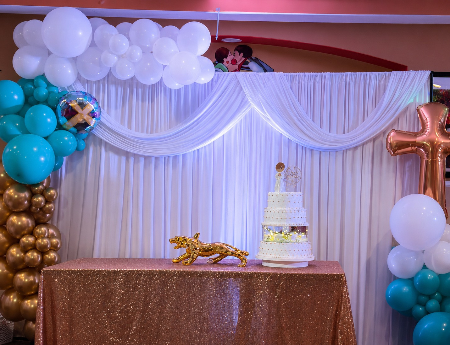 Decorated celebration backdrop with white draped curtains, white and teal balloons, a gold lion figure, a package-shaped cake with a topper, and a large rose gold cross balloon, set on a glittery table.