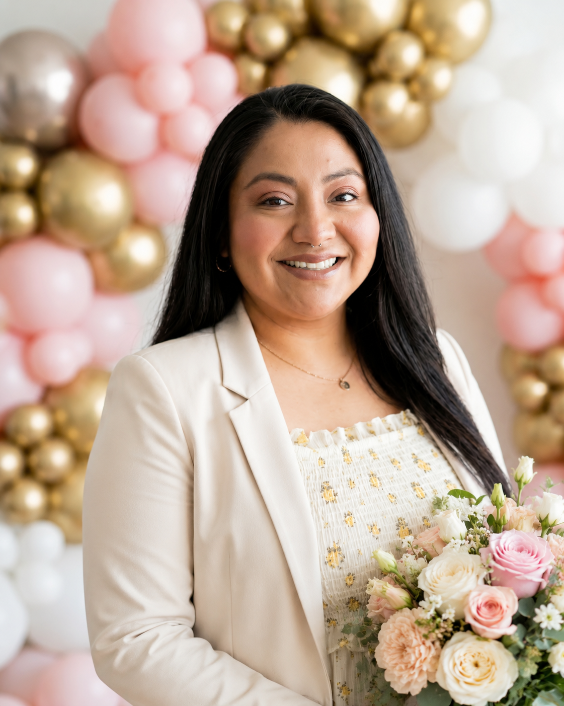 A woman with long black hair smiling, holding a bouquet of pink, cream, and white flowers. She is dressed in a light-colored blazer and patterned top. The background features a decorative balloon arrangement in pink, gold, and white.