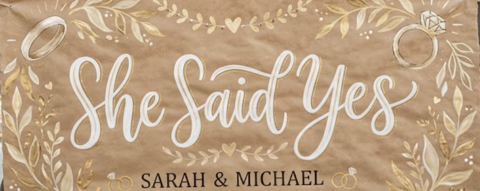 A decorative wedding sign with the words "She Said Yes" in large white script, topped with gold floral accents, featuring wedding rings and diamond icons, and below the names Sarah and Michael.