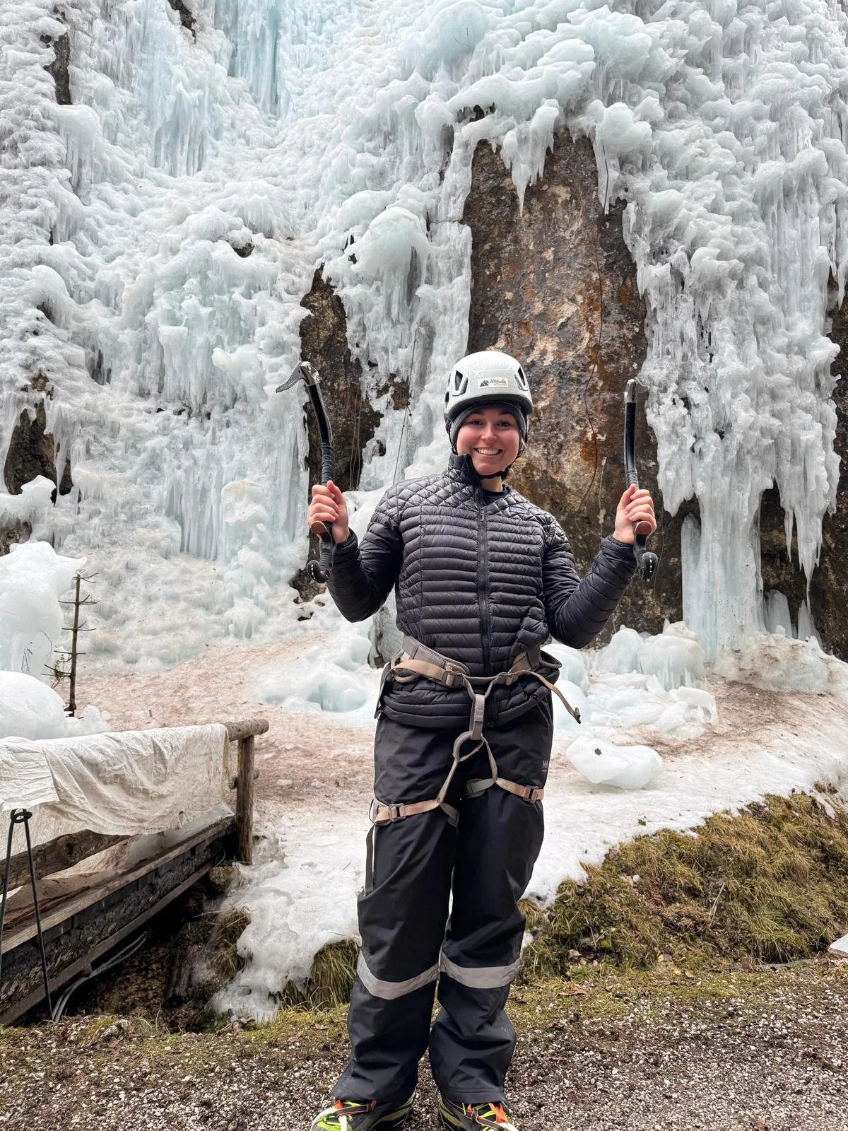 Person going on off trail adventures while wearing winter gear and helmet, holding ice axes, standing in front of an icy waterfall and snow-covered ground outdoors.