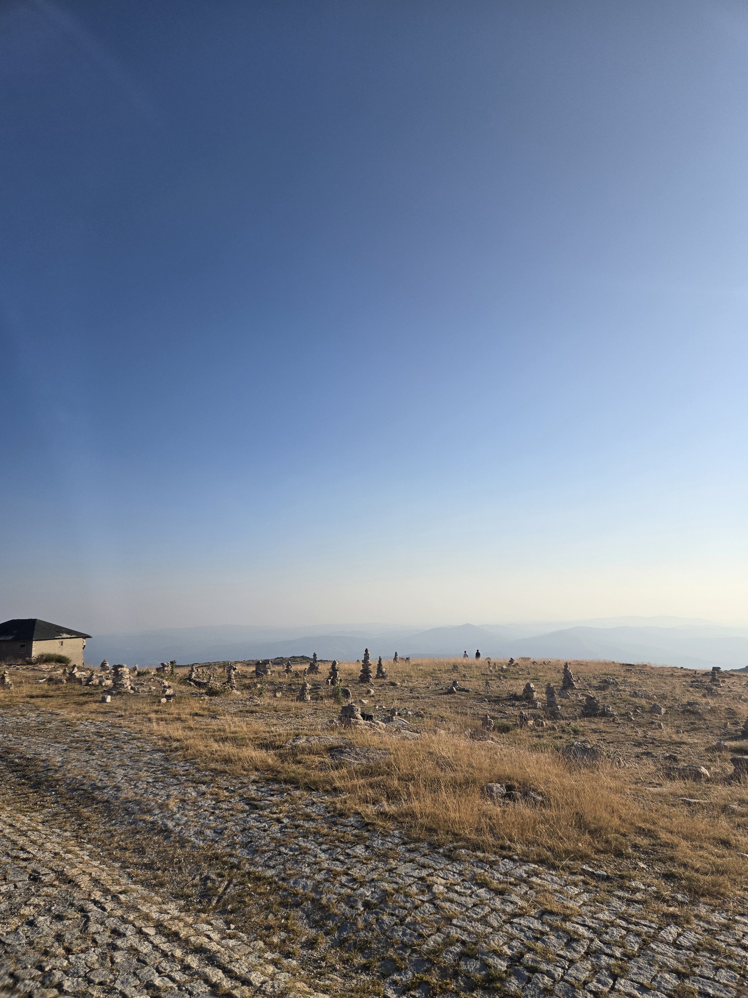 Wide view of a rocky, grassy plain with several stone cairns and a few scattered buildings, distant mountains under a clear blue sky, the perfect start to some off trail adventures.