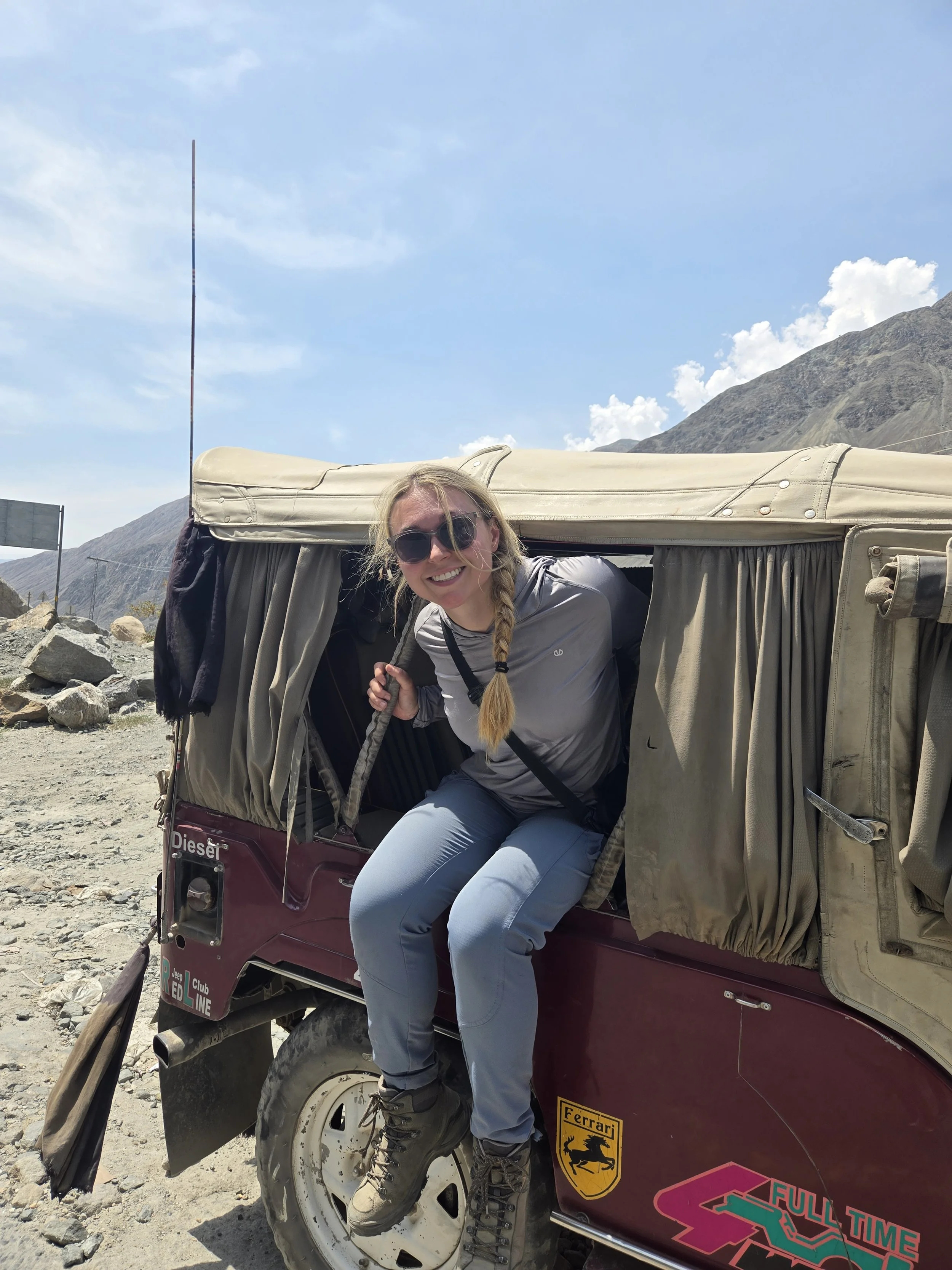 A smiling woman with long blonde hair, wearing sunglasses and outdoor clothing, is leaning out of a safari vehicle with beige canvas top, against a mountainous landscape with blue sky and clouds after a few days of off trail adventures.
