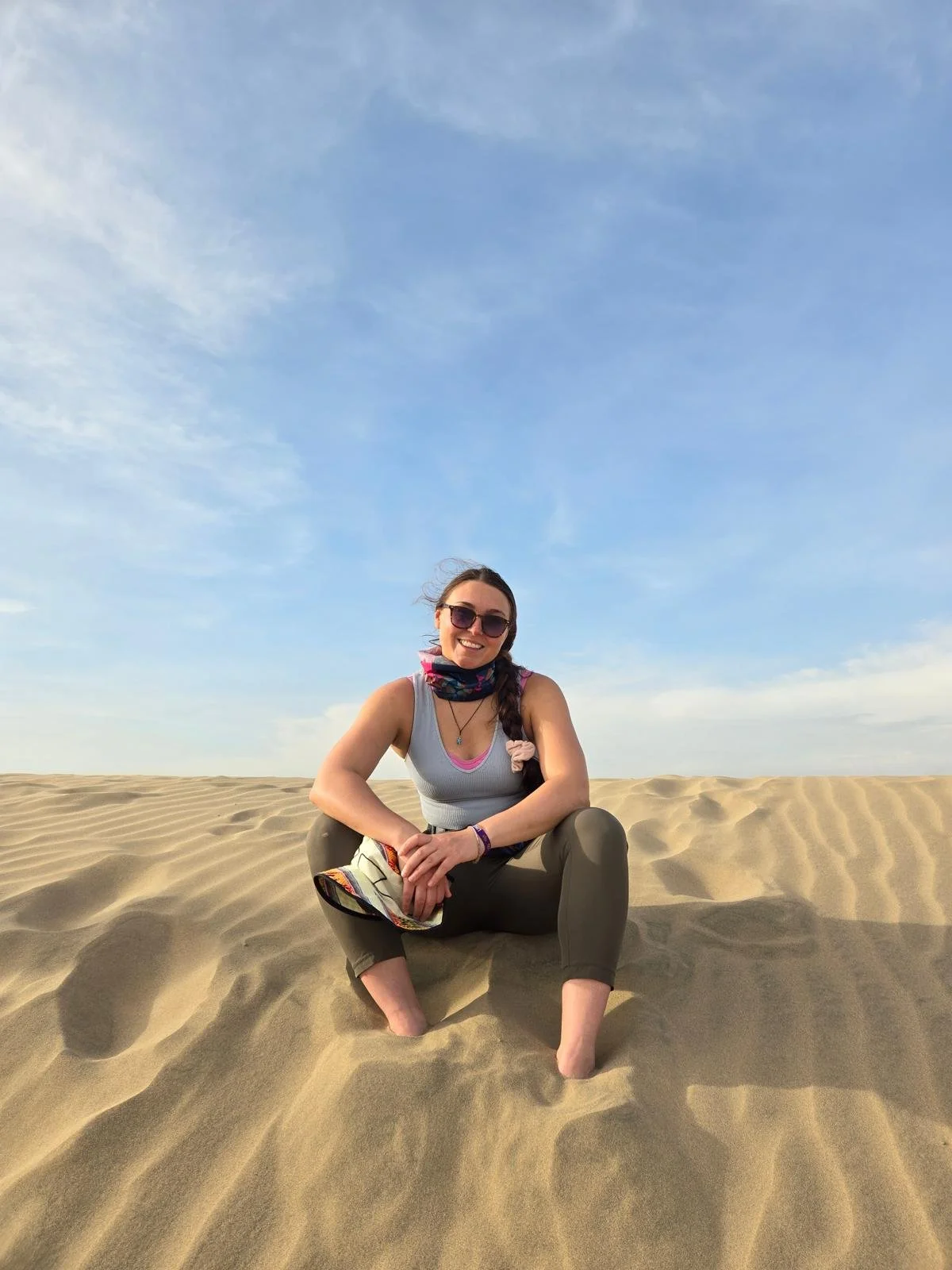 Woman with sunglasses, a scarf, and a tank top sitting cross-legged on sand dunes under a blue sky with scattered clouds, likely on a series of off trail adventures.