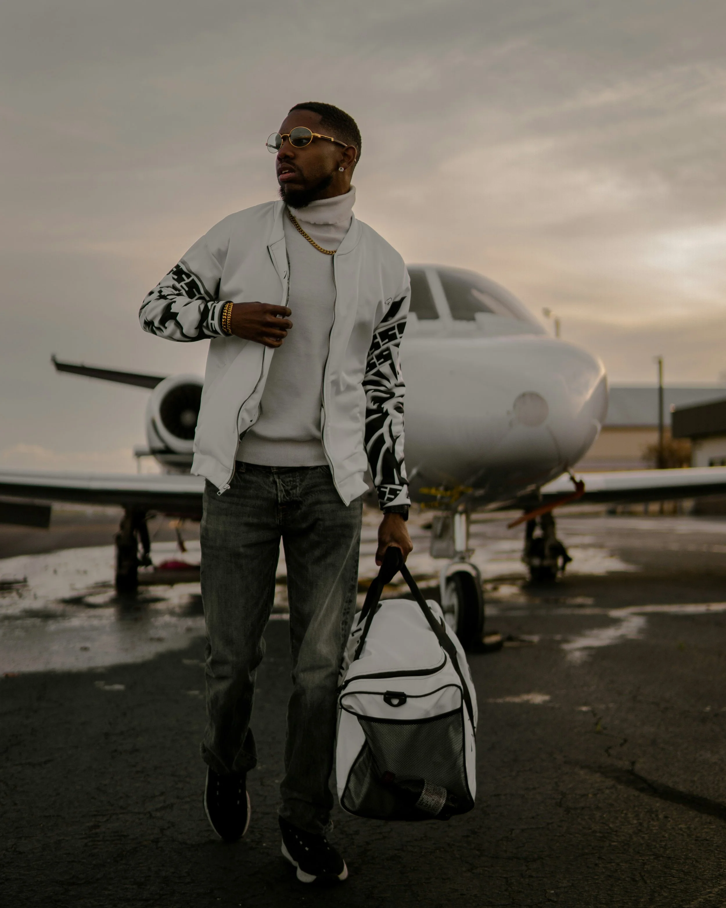 A man with sunglasses, jewelry, a white jacket, and gray pants, walking on an airport tarmac near a small private jet during sunset, carrying a duffle bag and pulling a travel bag with wheels.