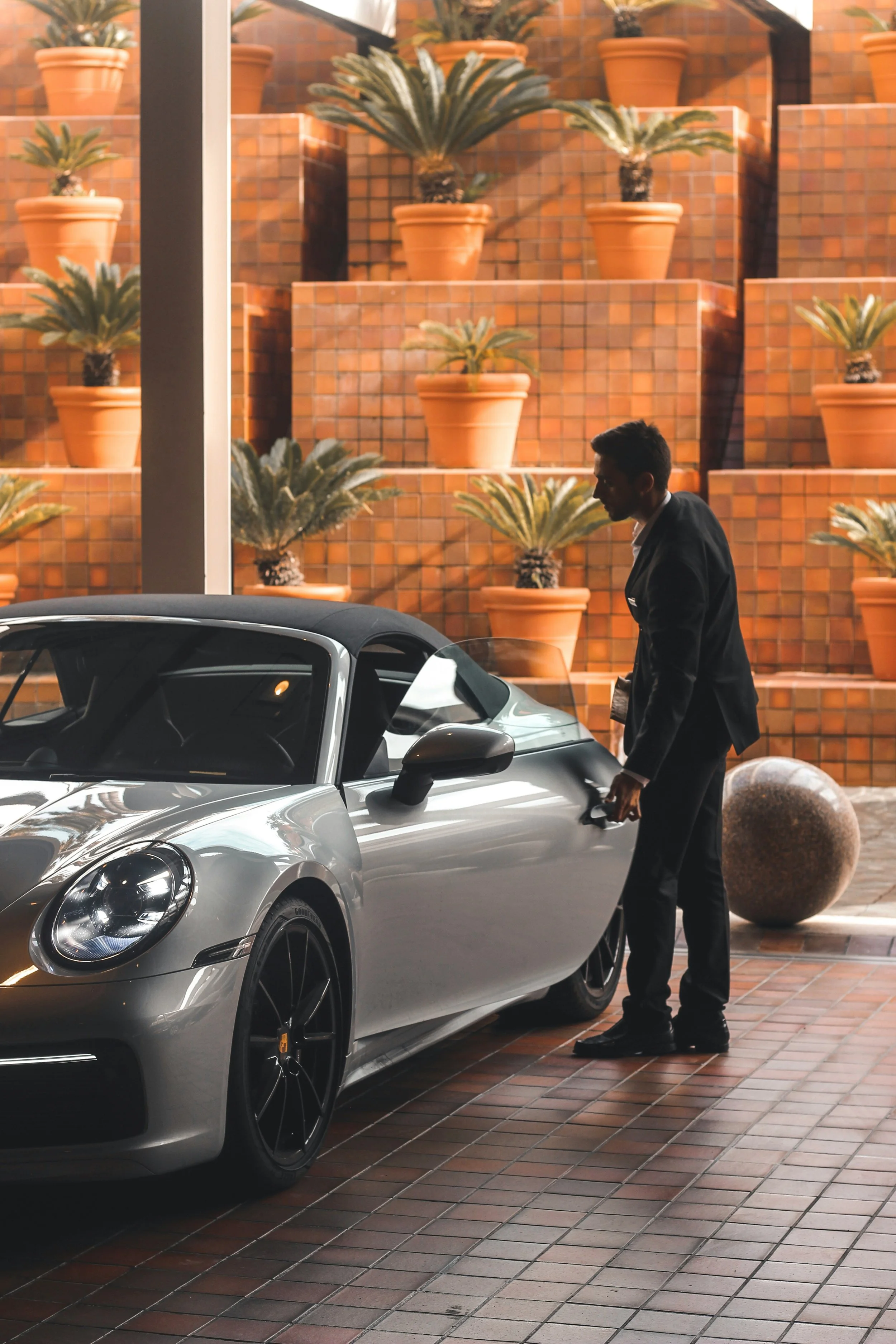 A man dressed in a black suit is standing next to a silver sports car with an open top, on a brick pavement. Behind him, there is a large decorative brick wall with terracotta pots containing palm plants and decorative stairs.