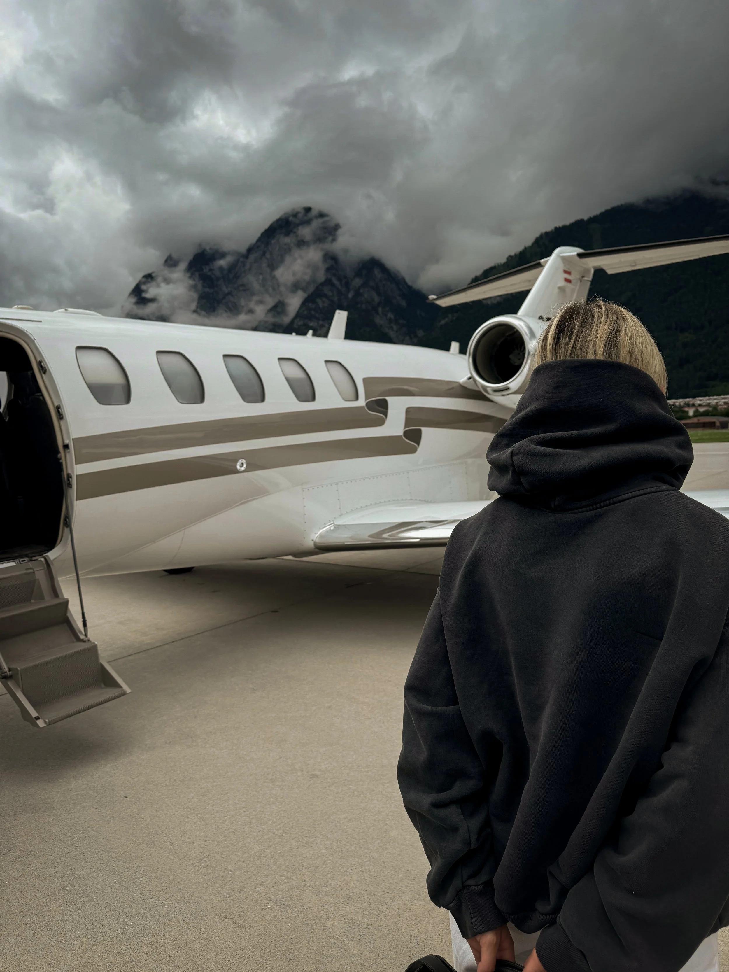 Person with blonde hair in a black hoodie standing near a private jet on an overcast day with mountains in the background.