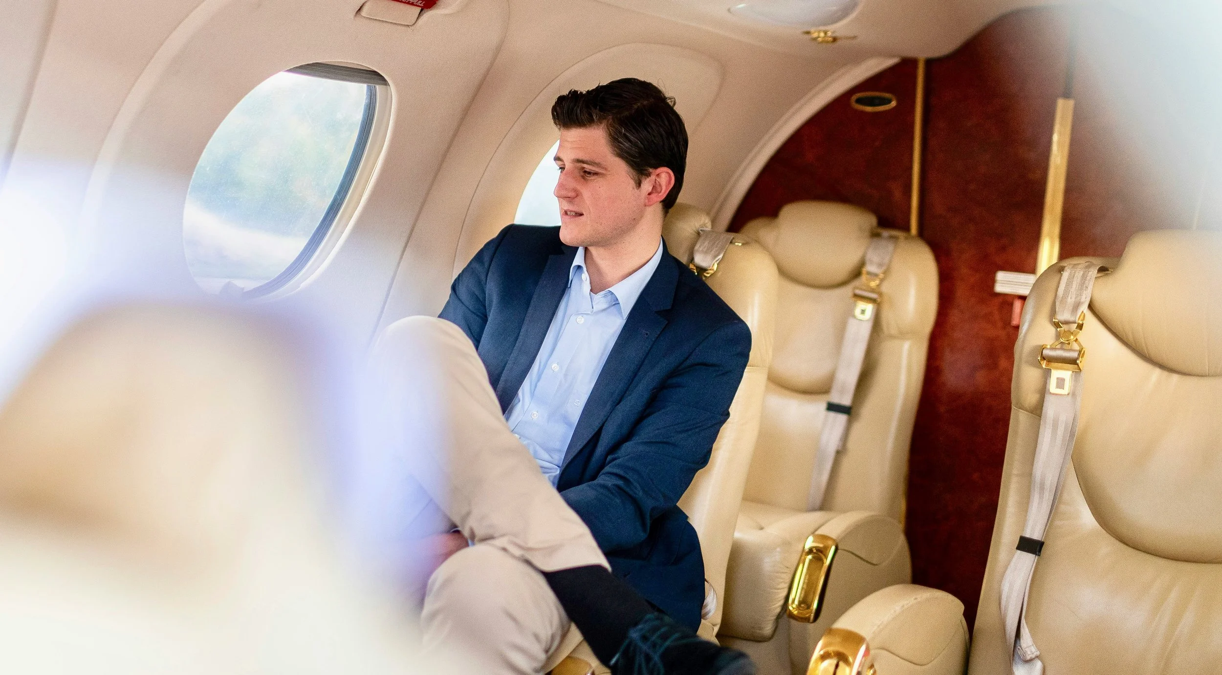 A man in a blue suit and light blue shirt sitting inside an airplane, looking down at something in his lap, with beige leather airplane seats around him.