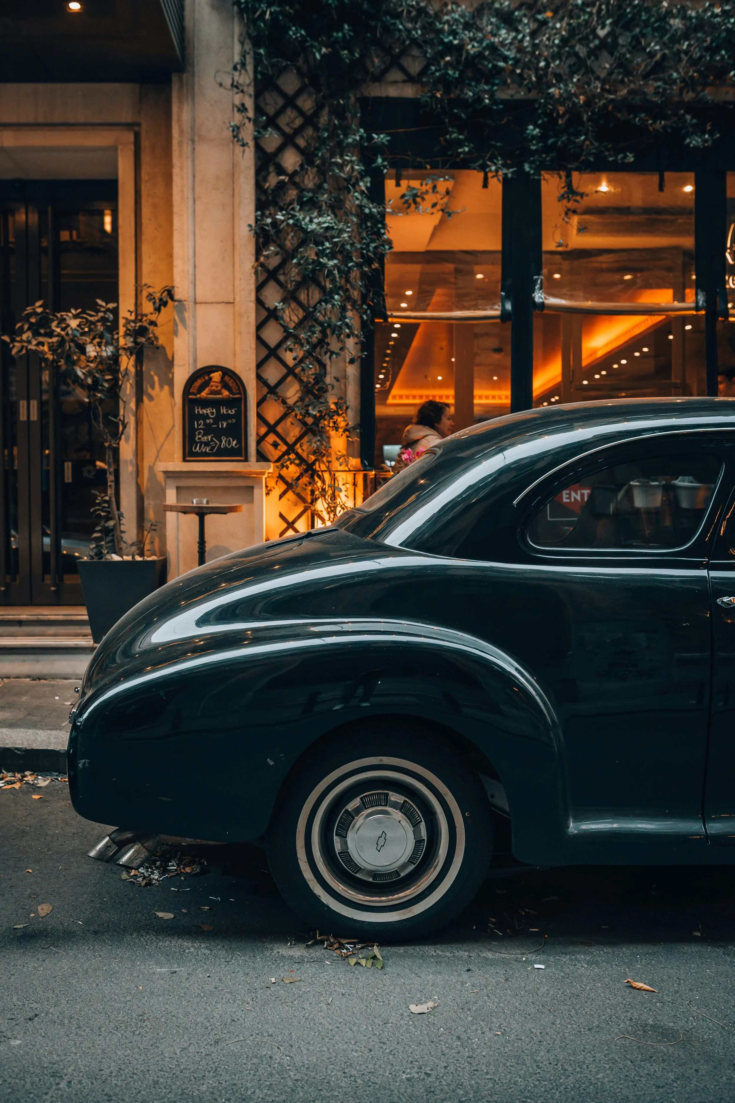 Close-up of a vintage black car parked on the street with a building and a window behind it during the evening. The street has some fallen leaves.